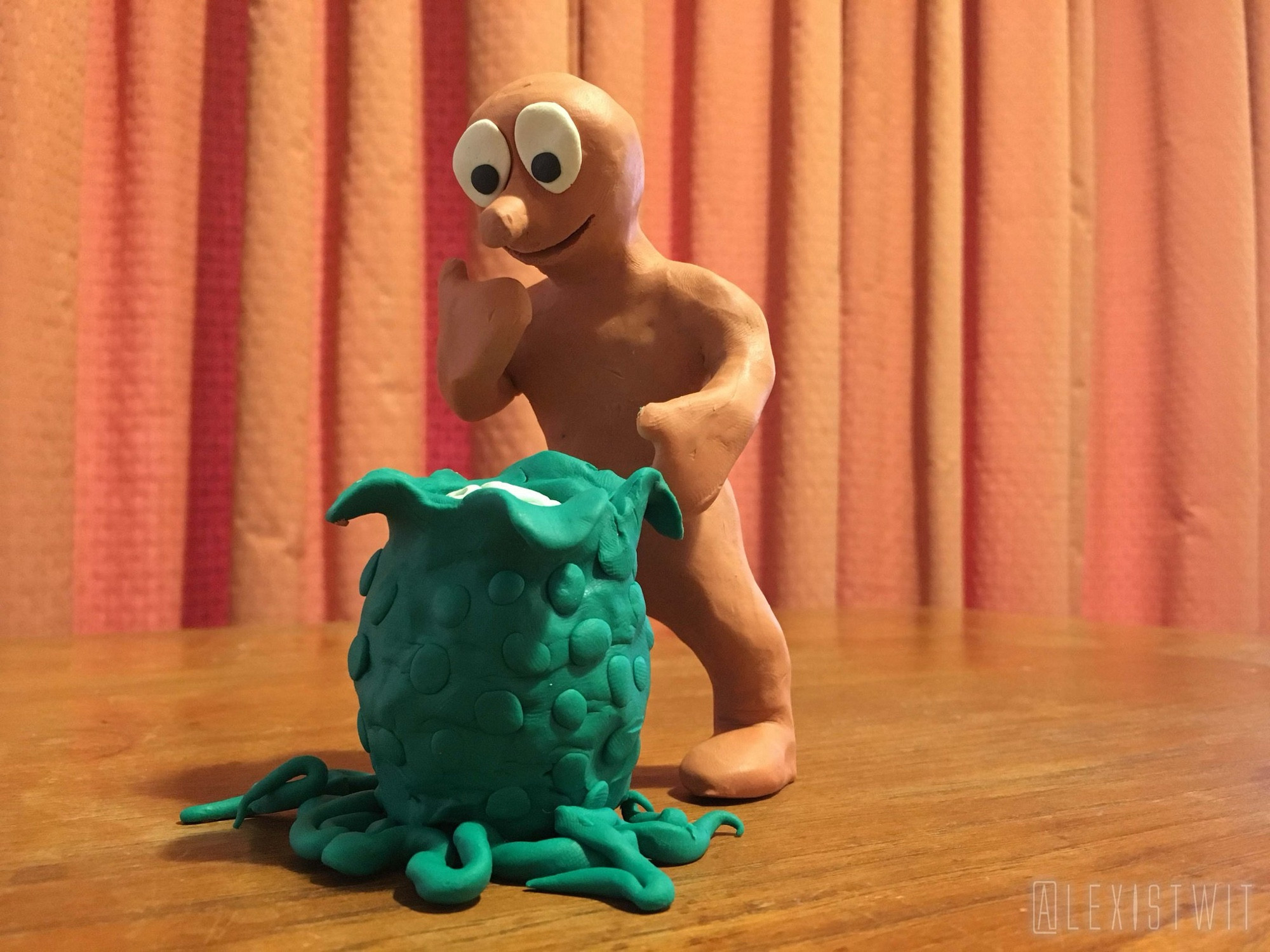 On a wooden tabletop, with a hideous orange curtain as a backdrop, the beloved claymation TV character Morph approaches a knobbly green plasticine alien egg, one hand raised to his mouth in an expression of curiosity as the petals on the top of the egg peel open to provide a glimpse of its pulsating white interior