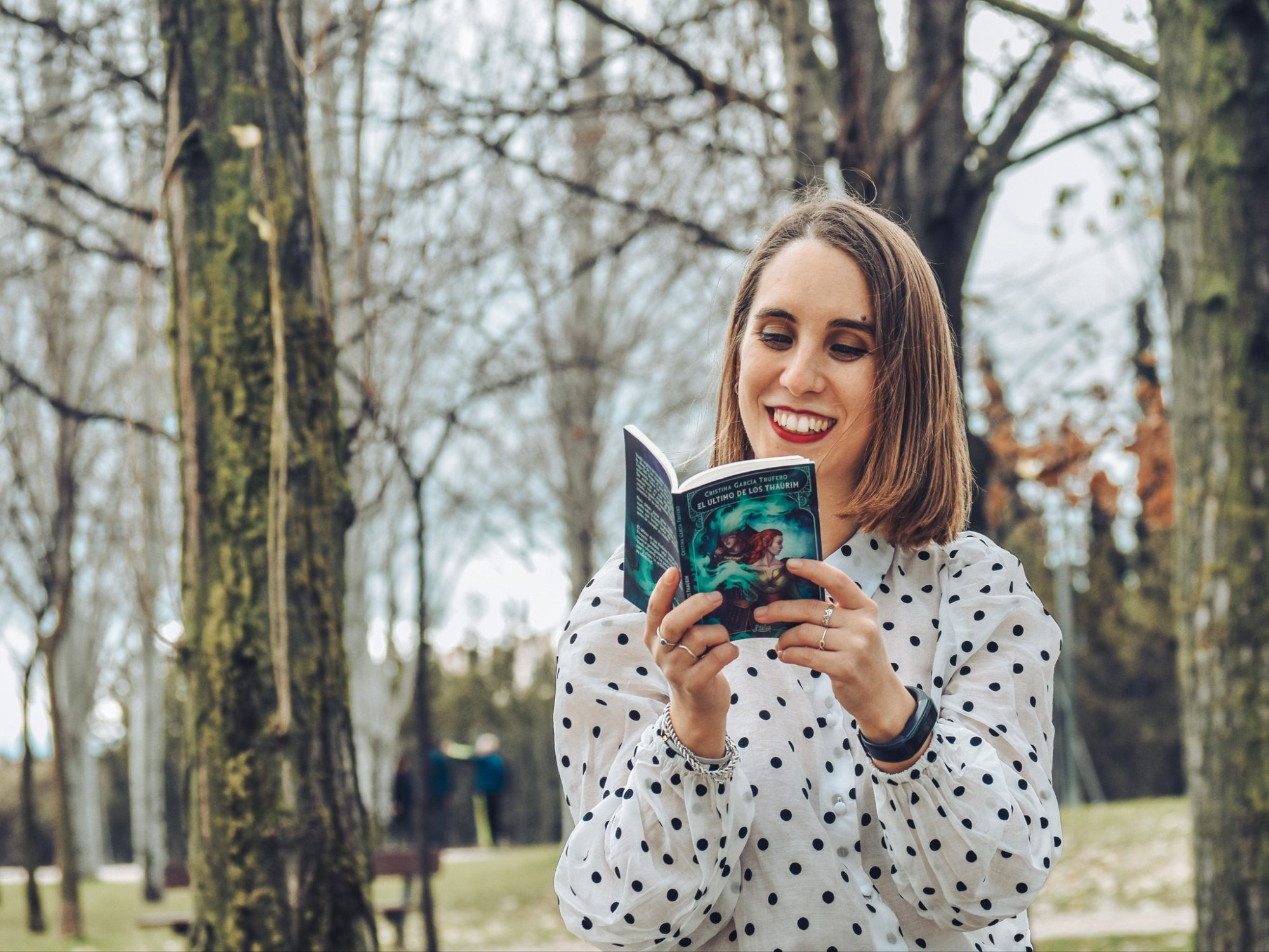 Fotografía en la que aparezco en un parque. Estoy leyendo mi novelette "El último de los thaûim" con una sonrisa en la cara. Llevo el pelo suelto y liso hasta los hombros, los labios rojos y una camisa de manga larga blanca con topos negros.