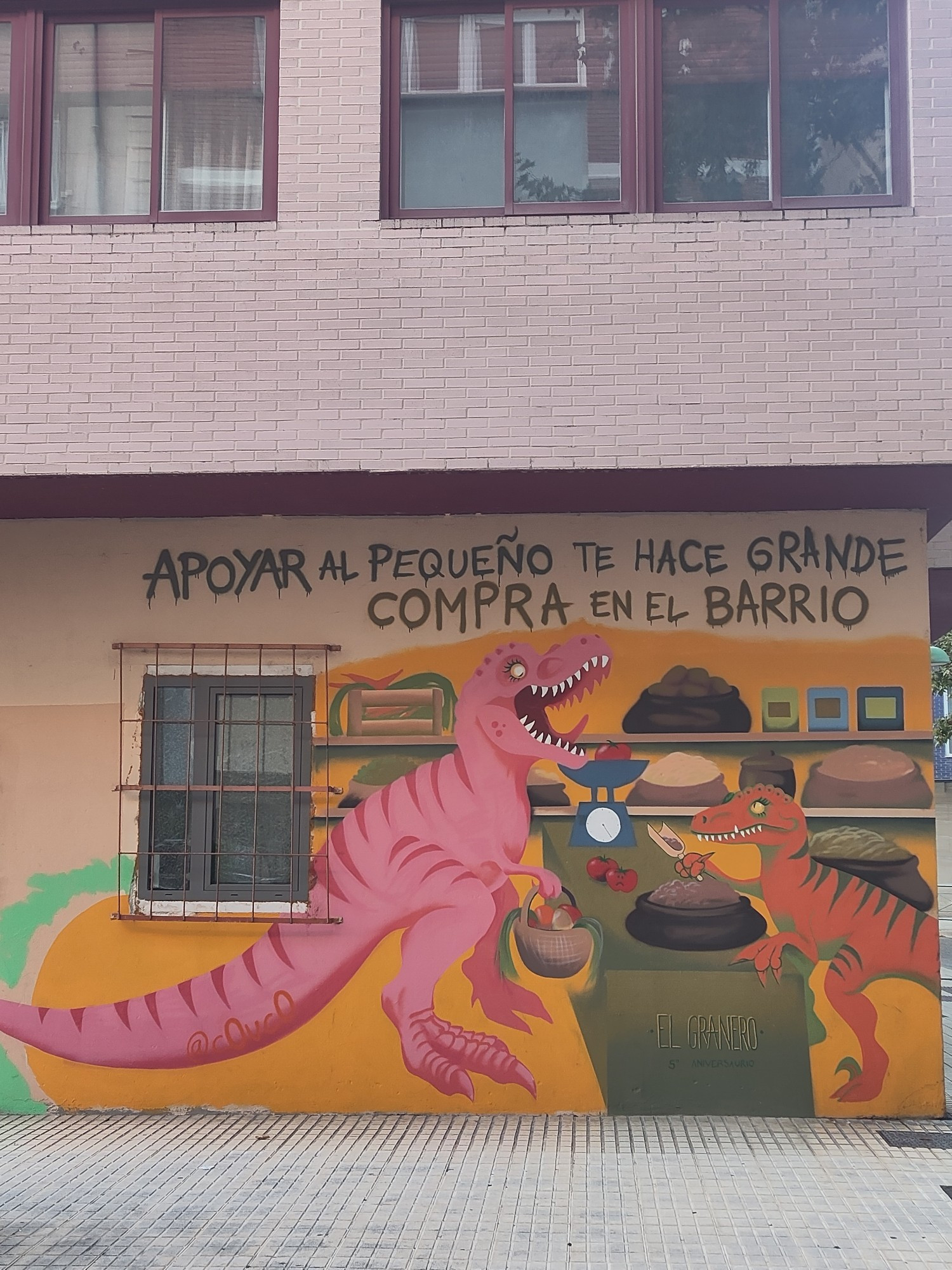 Mural con el texto «Apoyar al pequeño te hace grande. Compra en el barrio».  Al fondo hay dos baldas con cajas y sacos de productos. En primer término, un dinosaurio con una cesta con alimentos se encuentra ante un mostrador. En el lateral del mueble está escrito «El Granero. 5 Aniversario». Encima del mostrador hay una bascula antigua y dos tomates. Al otro lado otro dinosaurio está sacando con una pequeña pala producto de un saco.