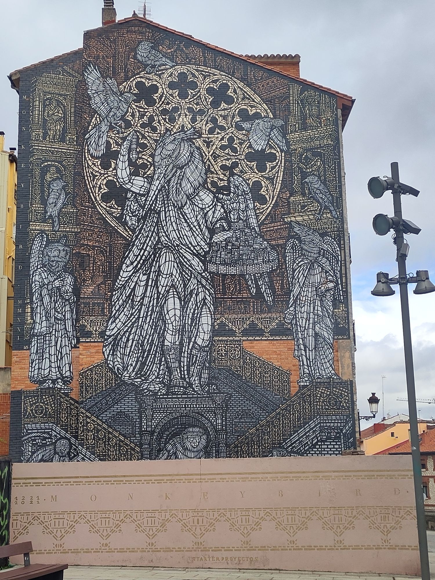 Mural que ocupa toda la medianera de un edificio. Al fondo, un retablo. En segundo término, el rosetón gótico de la puerta del Sarmental de la catedral de Burgos. En primer término, el Santo Ángel, patrono de la ciudad, sosteniendo en una mano una maqueta de la ciudad. Su cabeza es de paloma. Sobre el ángel vuelan palomas. En la parte inferior, la Escalera Dorada de la catedral.