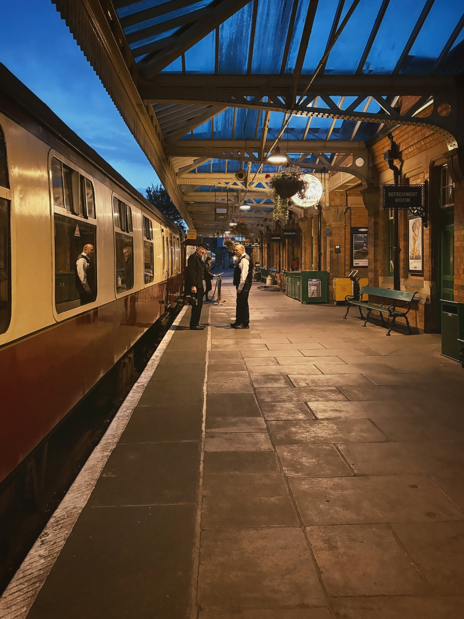 looking down the platform with steam train on the left and restored waiting rooms to the right. 2 train guards chat, reflecting in train windows. both are dressed in vintage uniforms and one holds an old lamp.