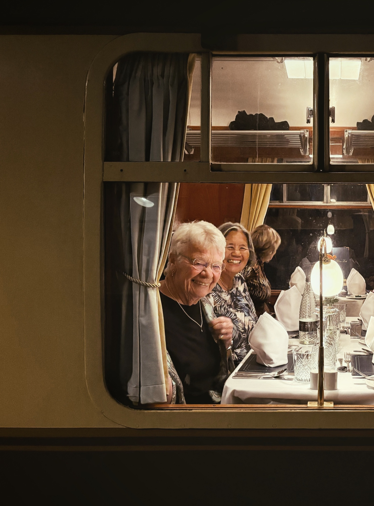 looking through a window framed with drawn curtains into the steam train. my smiling aunties are sitting at our table, which is laid with linens and ornate glassware. behind, a woman looks out the far window.