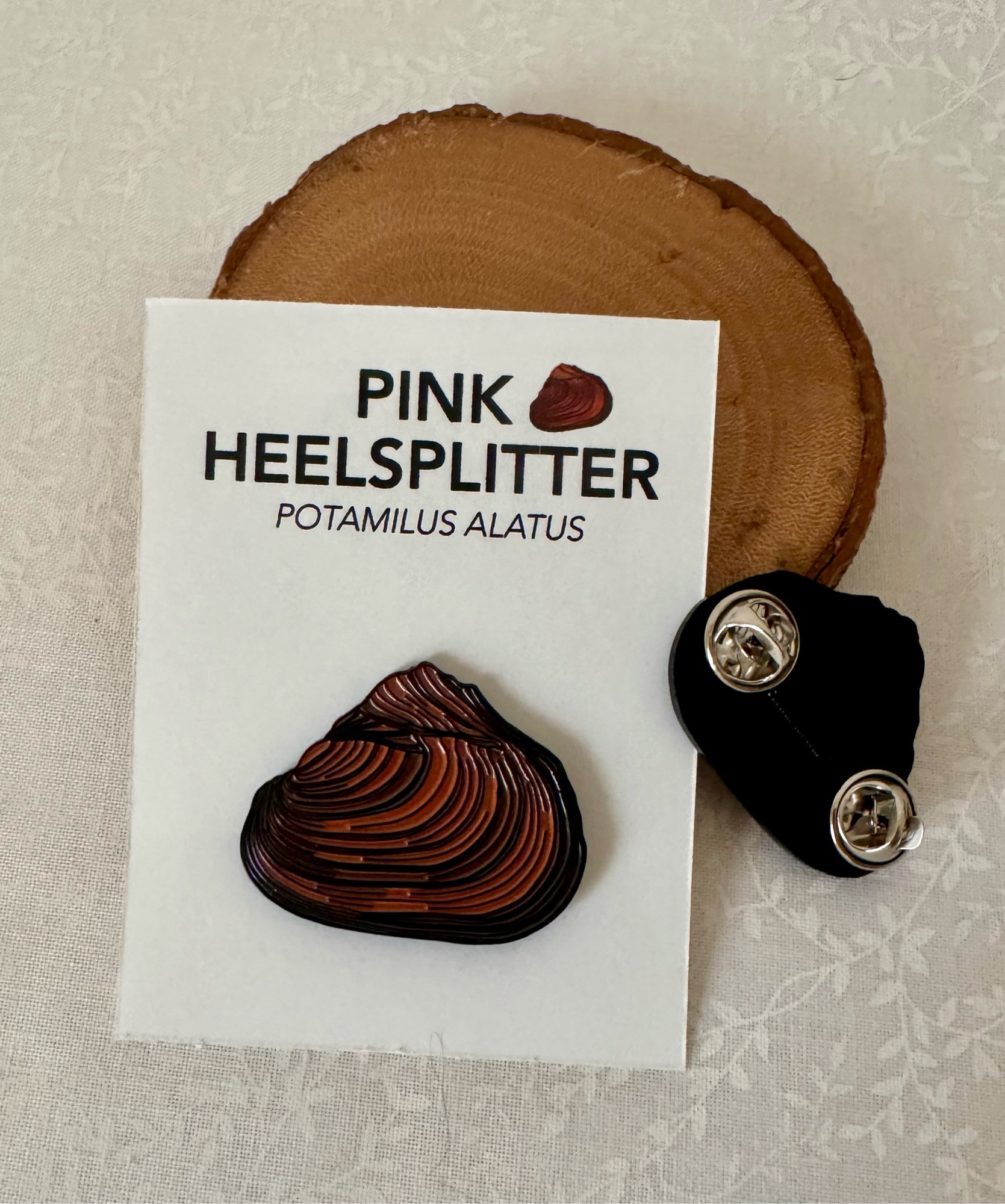 Pink heelsplitter mussel enamel pin (Potamilus alatus) displayed on a white backing card with a wood slice and pin backing visible. Ideal for aquatic biology enthusiasts and collectors of nature-themed accessories