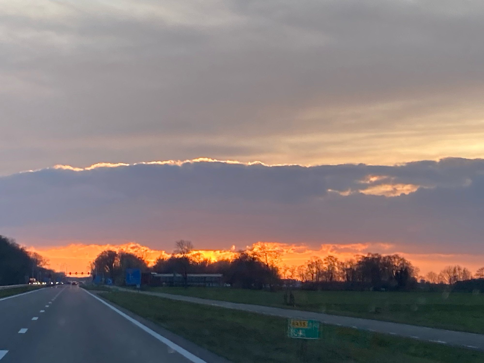 Vanuit de auto, snelweg, berm, bomenrij en avondzon met daarboven enige bewolking 