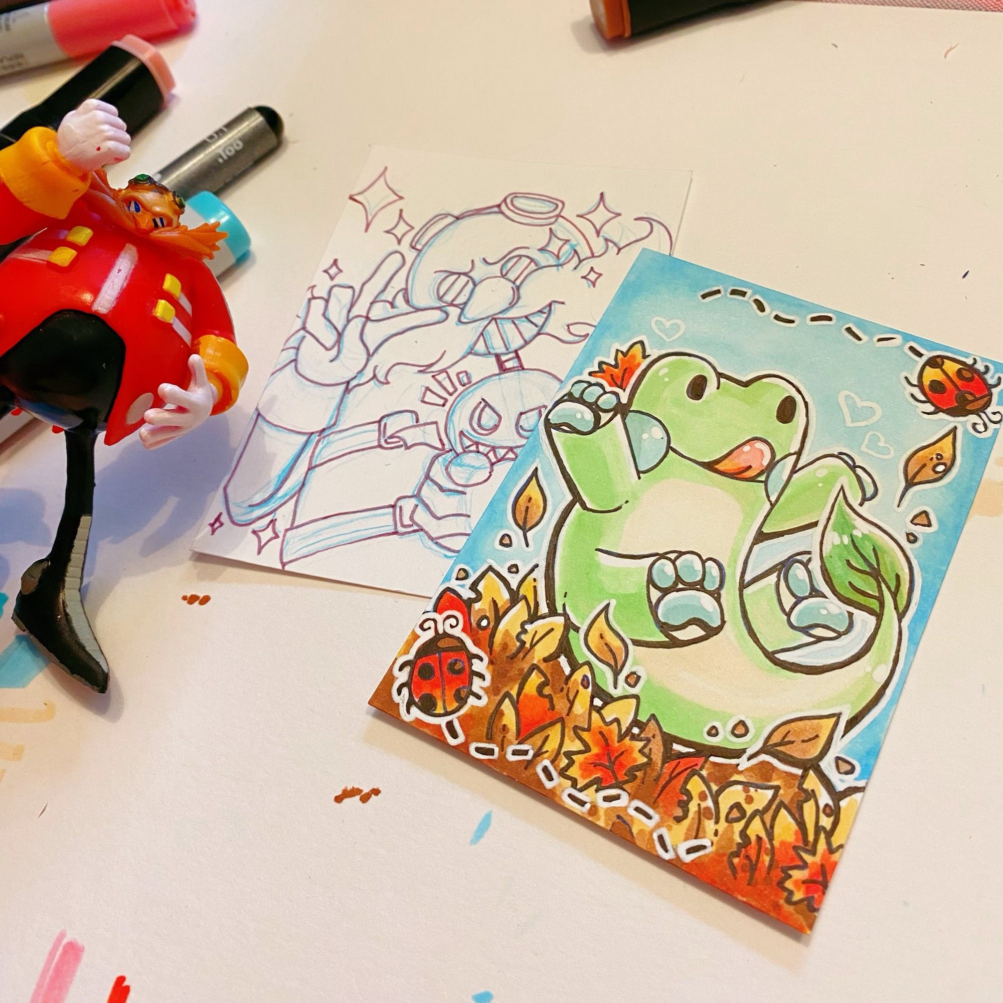 Traditional art (comic marker) of my mascot Plort, a frog/dog/plant hybrid jumping in a pile of autumn leaves. Also pictured is an unfinished art of Eggman from the Sonic franchise, and a little figure of Eggman peeking into the frame.