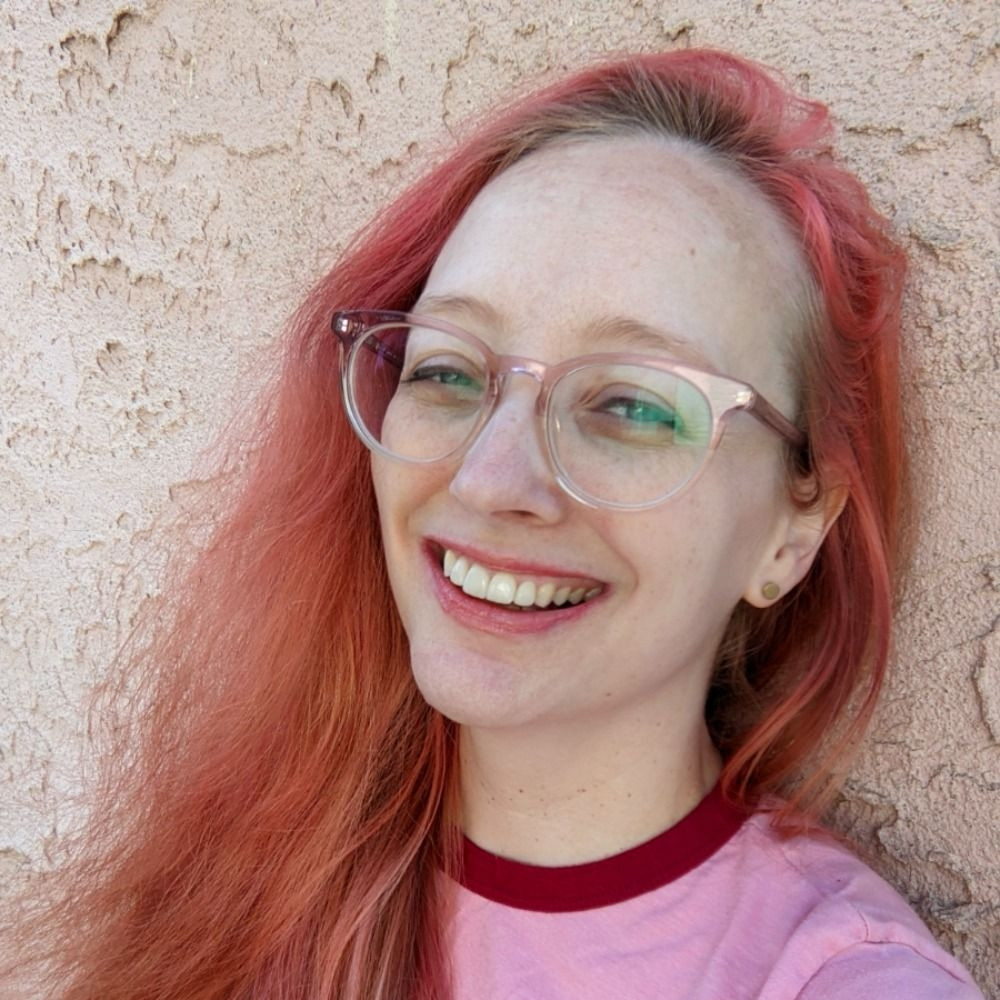 Here is Kali, a white woman with pink hair and pink glasses wearing a pink shirt in front of a pink wall. They're all different pinks.