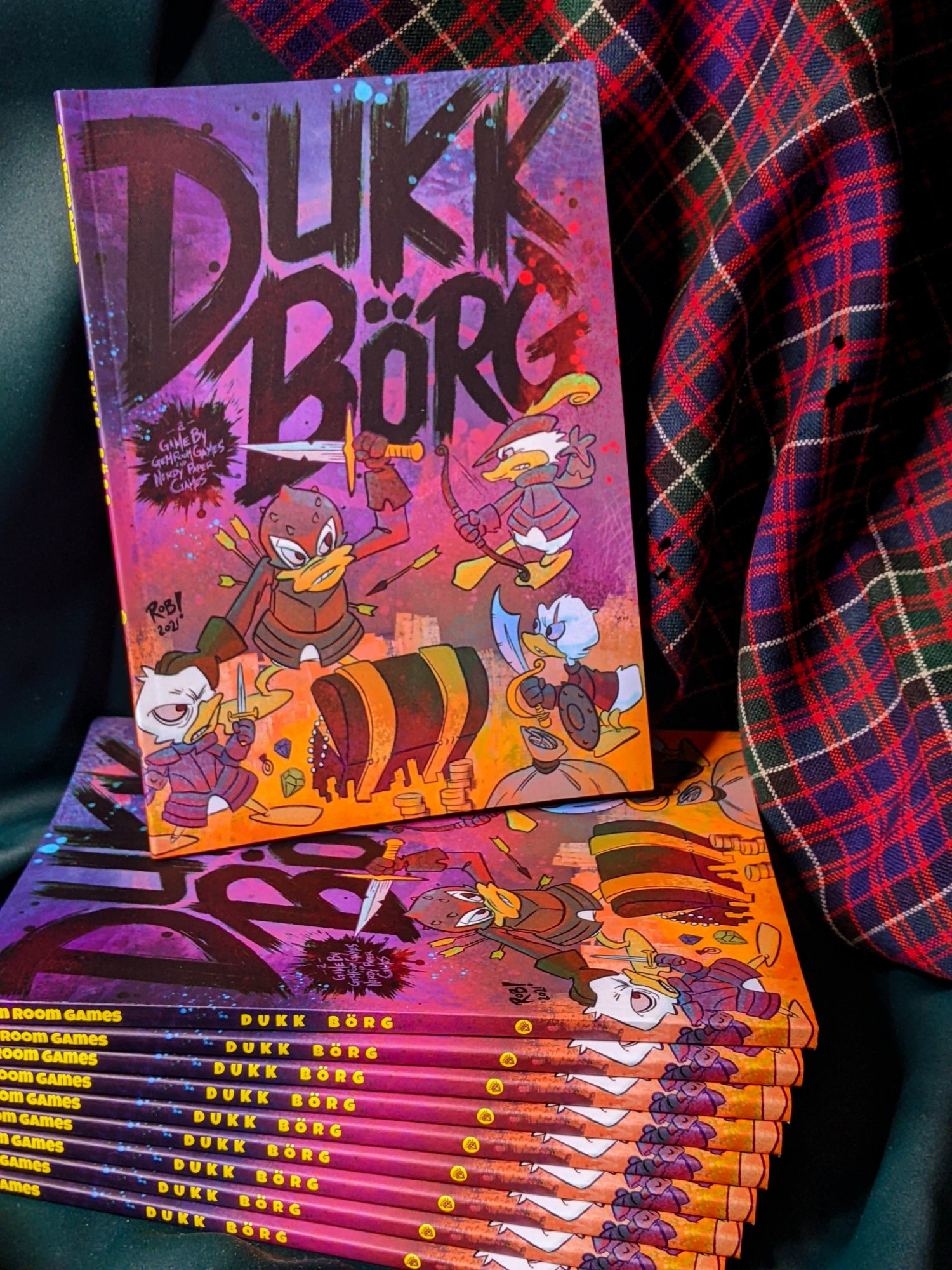 Stacked copies of DUKK BÖRG in front of a moth eaten tartan. The cover shows the title of the paperback book in scratchy dark letters above a band of cartoon adventurer ducks turning on each other for treasure.