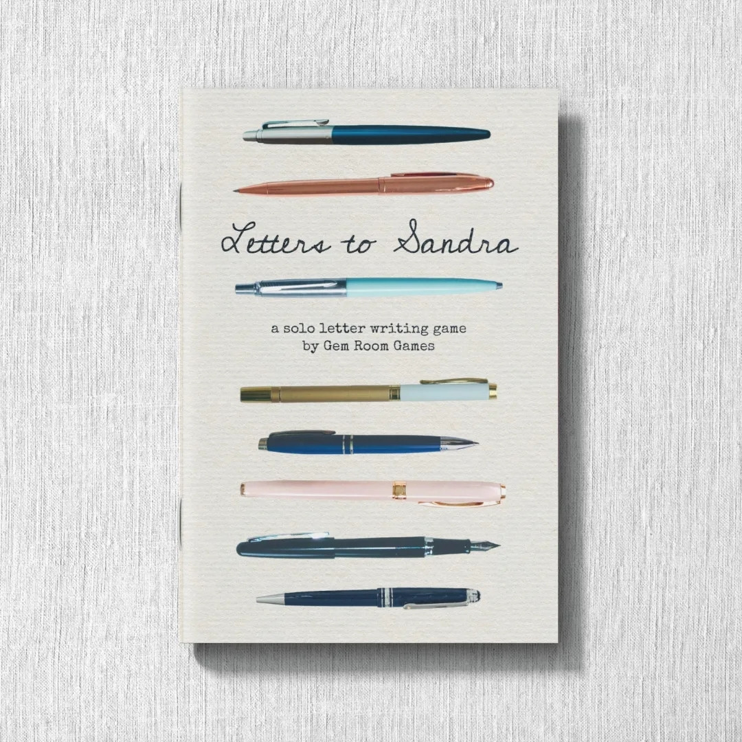 A single copy of Letters to Sandra on a pale textile background. The cover of the cream zine features the handwritten-looking title between colorful pens arrayed horizontally. Small typed text reads "a solo letter writing game by Gem Room Games."