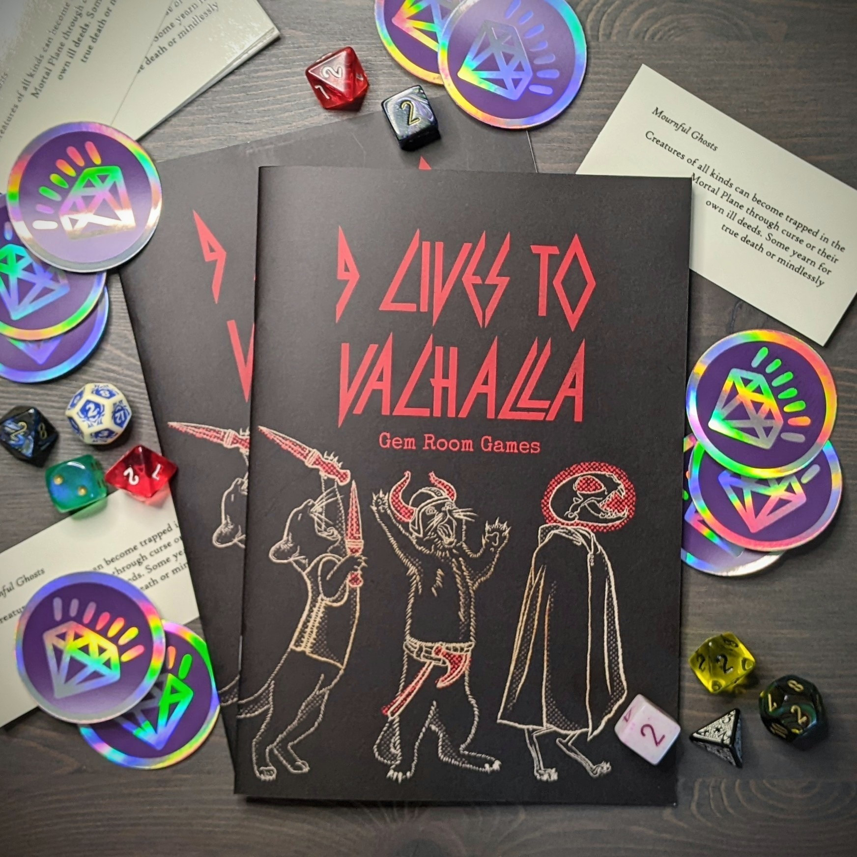 Two copies of 9 Lives to Valhalla sit on gray wood, surrounded by a scattering of holographic gem stickers and assorted dice. The covers feature white line illustration of yowling warrior cats following a cloaked skeleton cat beneath red text of the title. The font evokes retro heavy metal.