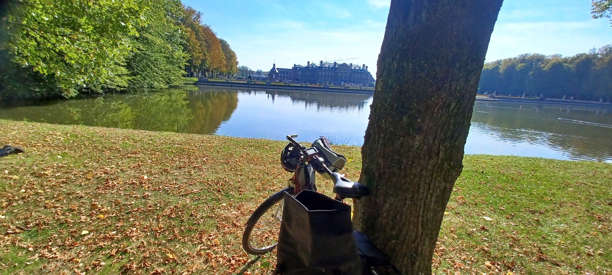 Ein Foto vom Schloss Nordkirchen, davor der glitzernde Schlossteich, im Vordergrund ein rotes Treckingrad an einen Baum gelehnt und eine sonnige Wiese mit Herbstlaub.