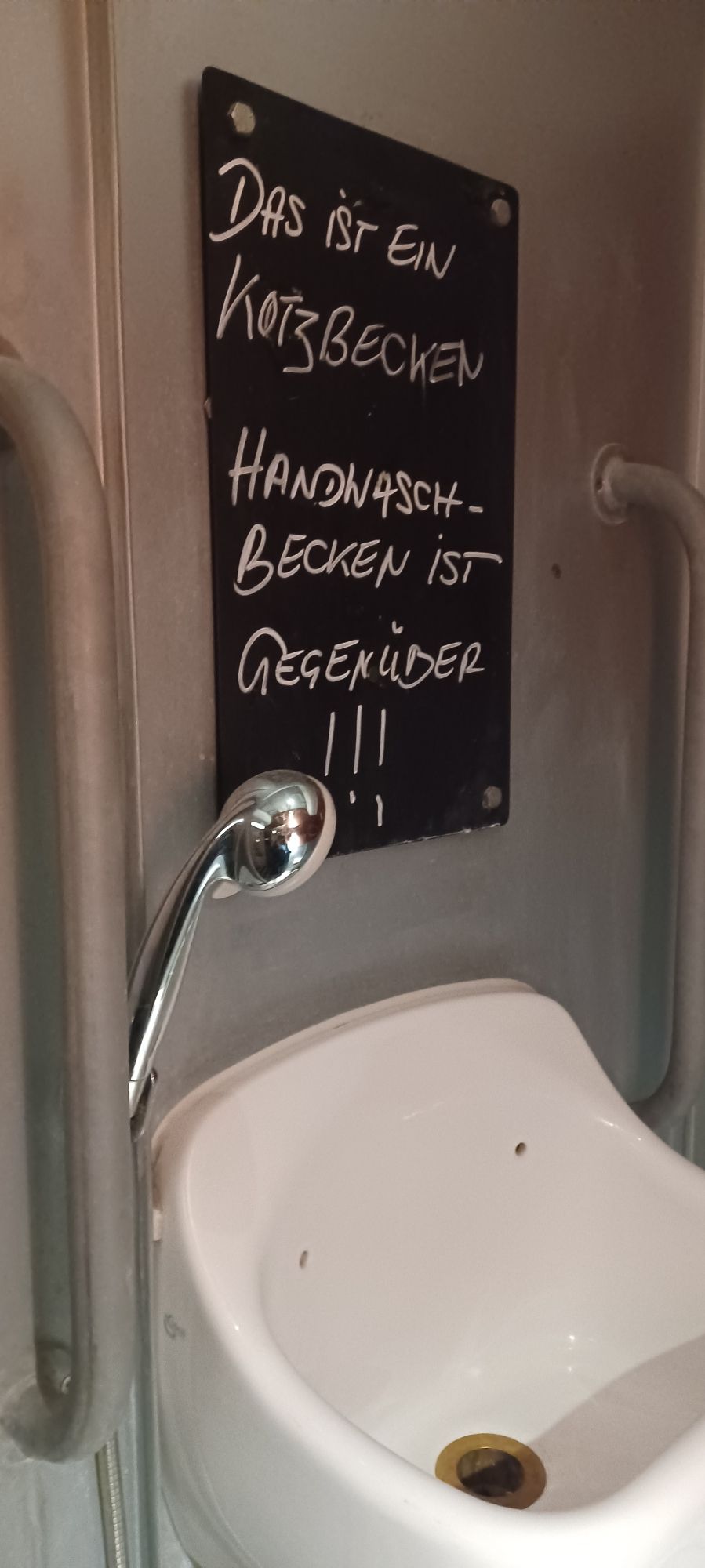 Waschbecken mit Duschschlauch.Darüber der Satz: Das ist ein Kotzbecken, Händewaschen gegenüber.