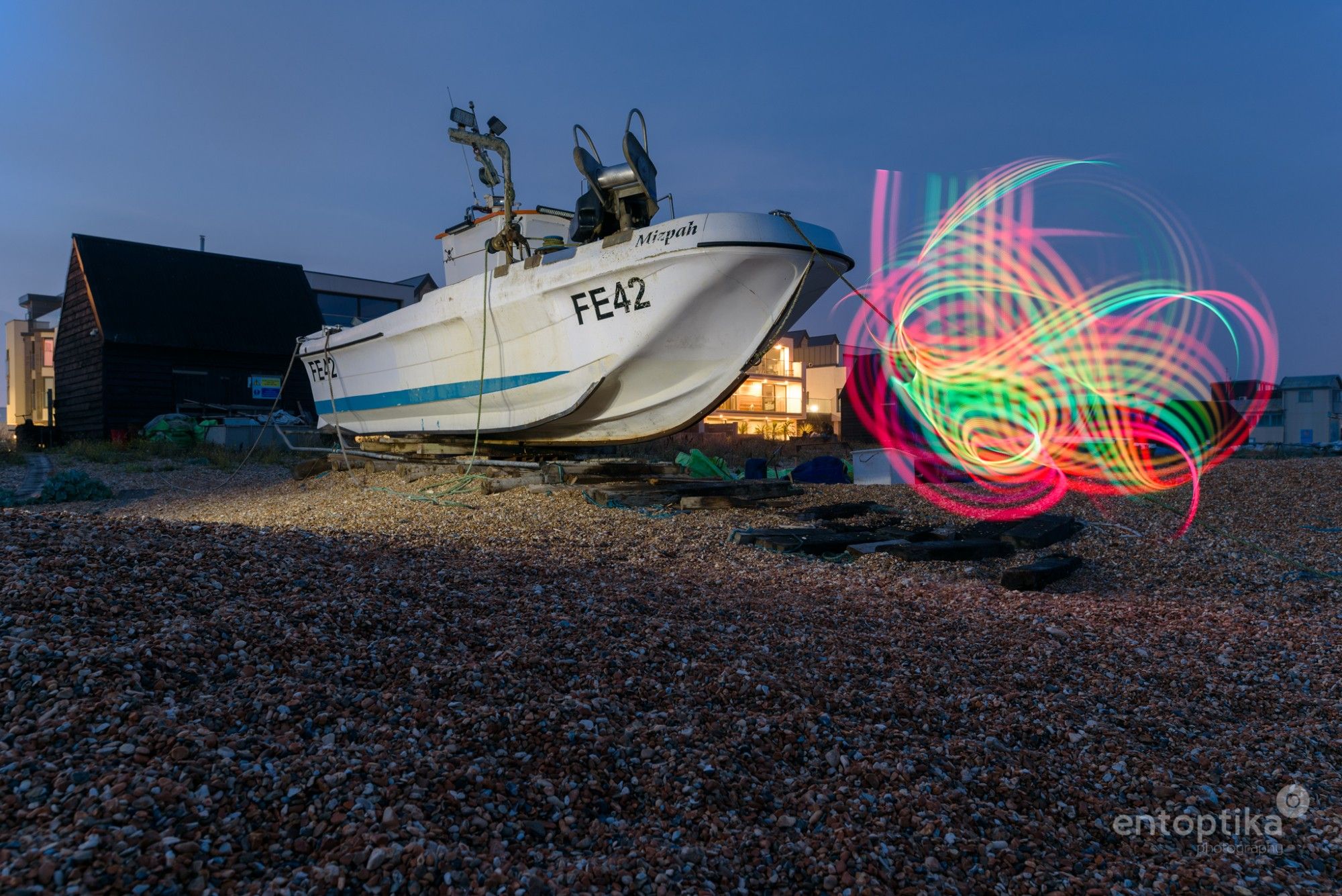 An evening image of a boat on the shingle beach of Hythe in Kent, with light trails in front of it.