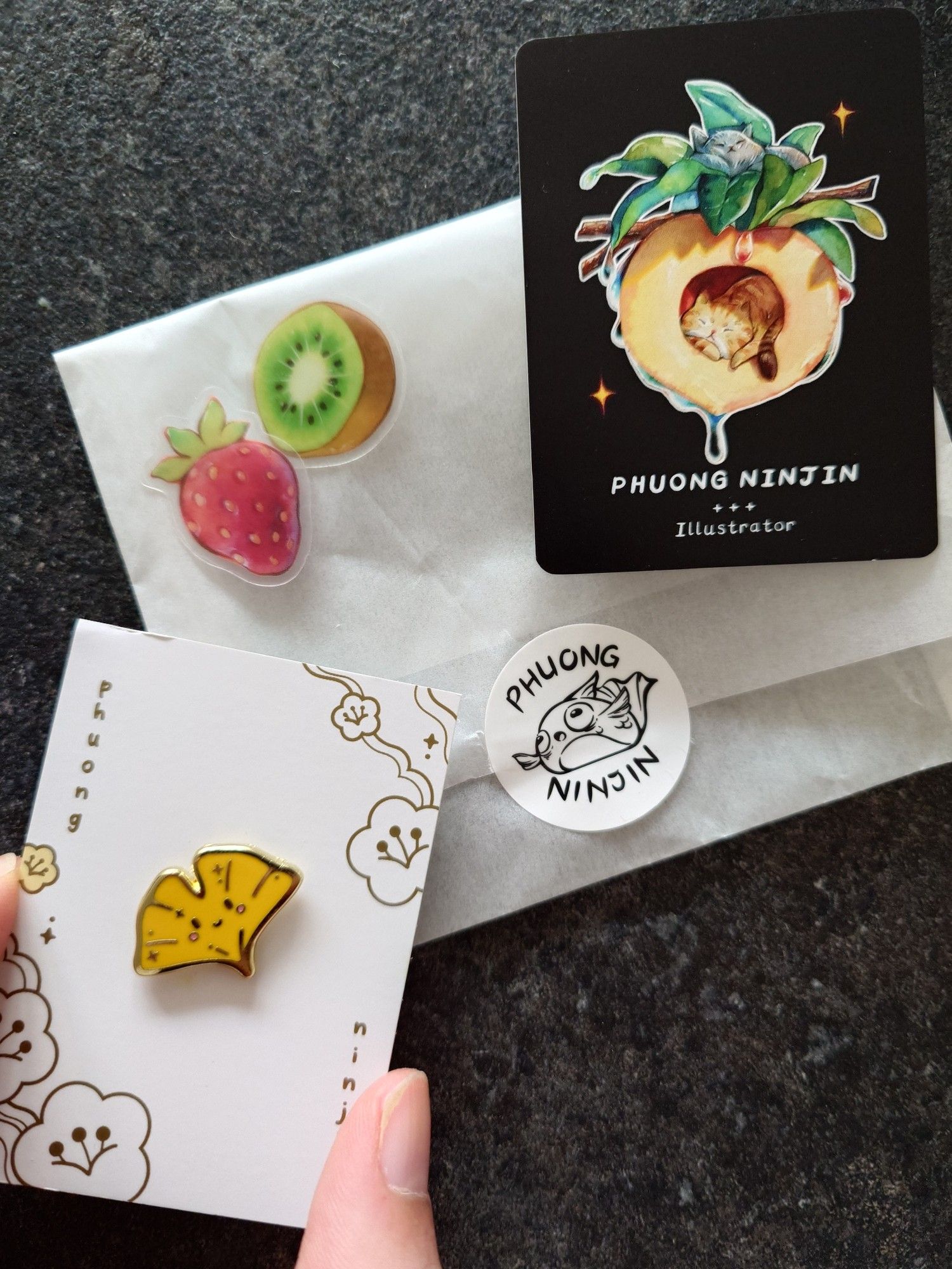 Artist merch purchased: a smiley ginko leaf pin, the artist business card - Phuong Ninjin, and two free bonus stickers of a strawberry and kiwi.