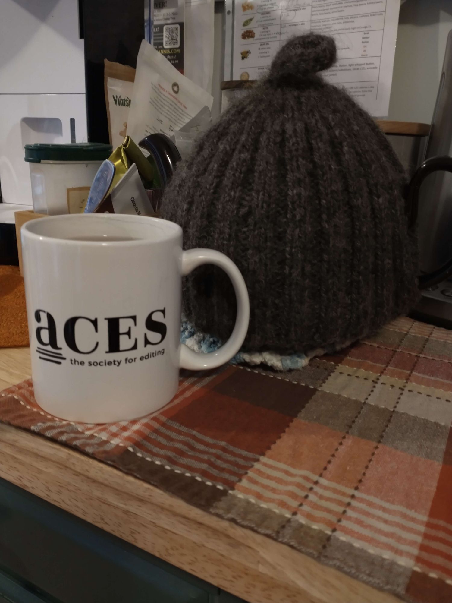 White mug with "ACES: the society for editing" printed on it, sitting on a plaid cloth. Next to it is a dark grey knitted tea cozy. Kitchen items and papers visible in the background.