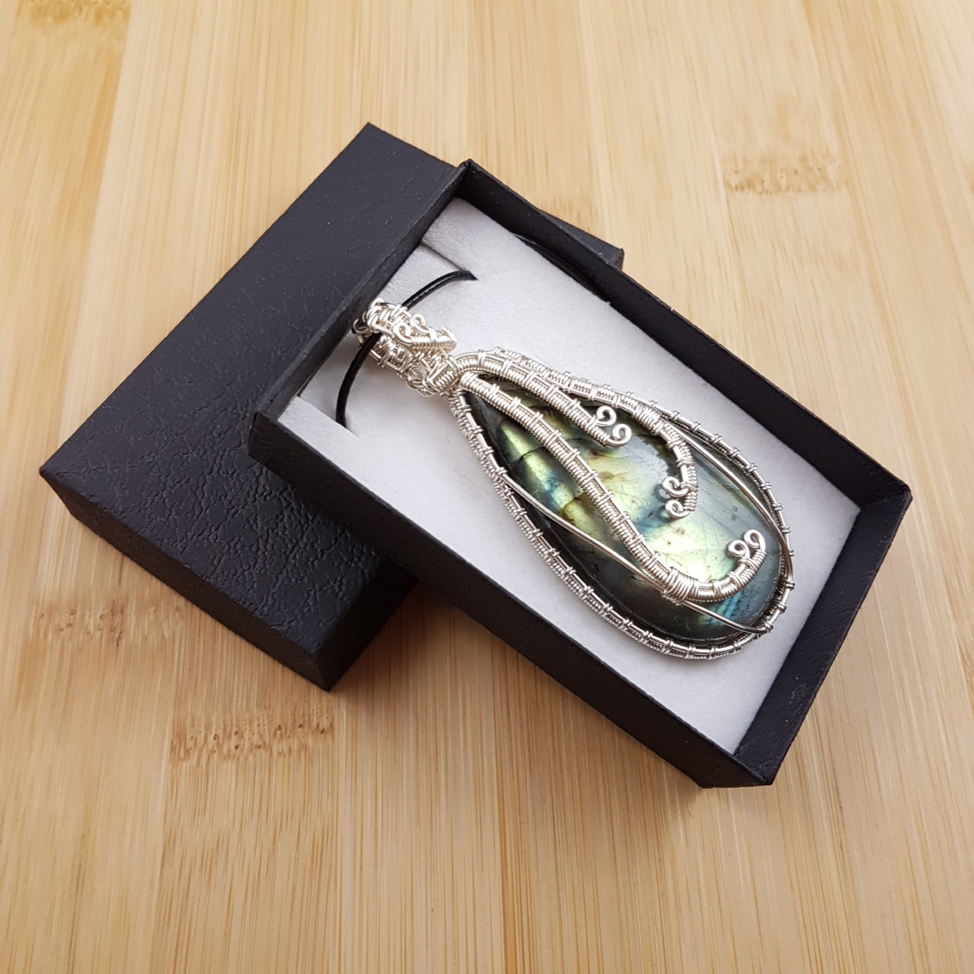 Large glowing green drop pendant wrapped in wavy silver wire, sitting in black and white gift box.