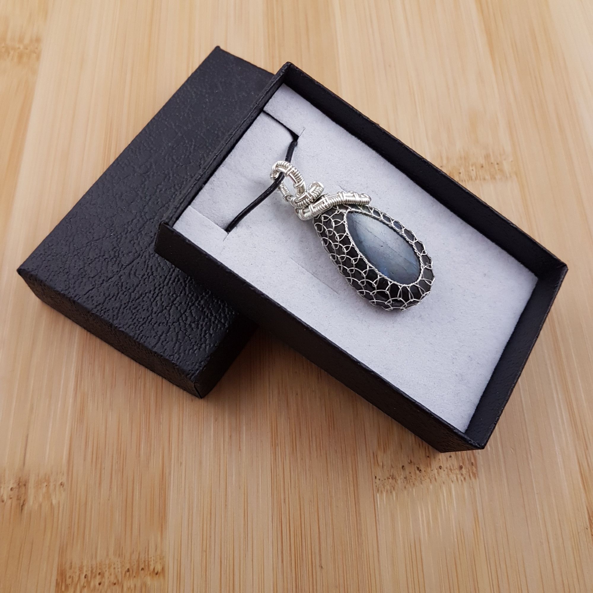 Small glowing blue oval pendant netted in silver wire, sitting in black and white gift box.