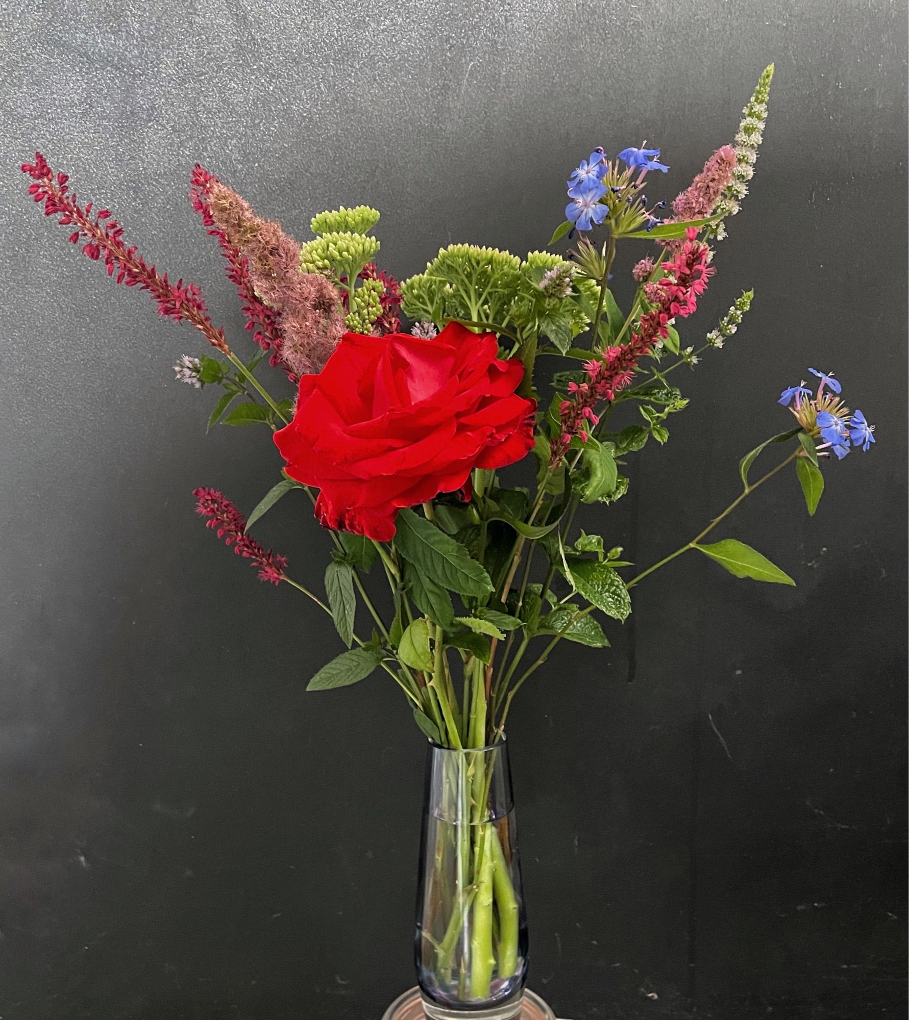 Flower arrangement - one large deep red rose surrounded by spikes flowers in pinks and purples, plus pointed leaf foliage and pale green ice plants, all against a dark chalkboard background.