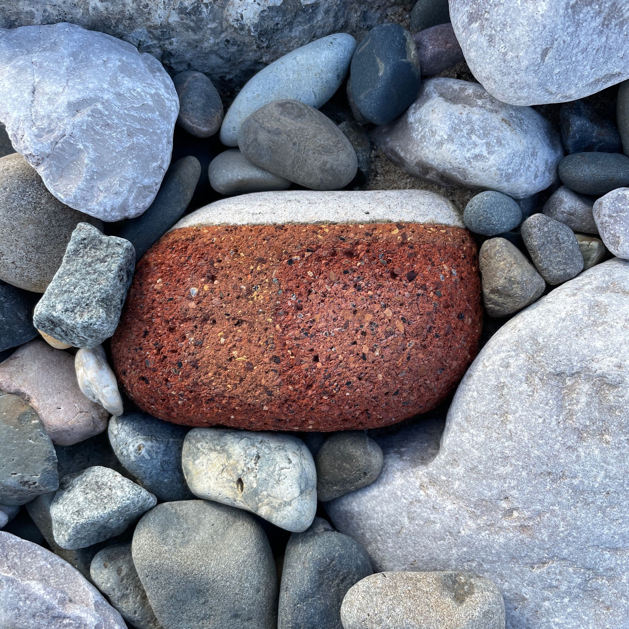 A roughly square piece of sea worn brick, red and dappled with holes with a layer of pale mortar on the top that looks like icing. It rests on blue grey pebbles and rocks which are a contrasting colour and make the cake was of the brick more obvious.