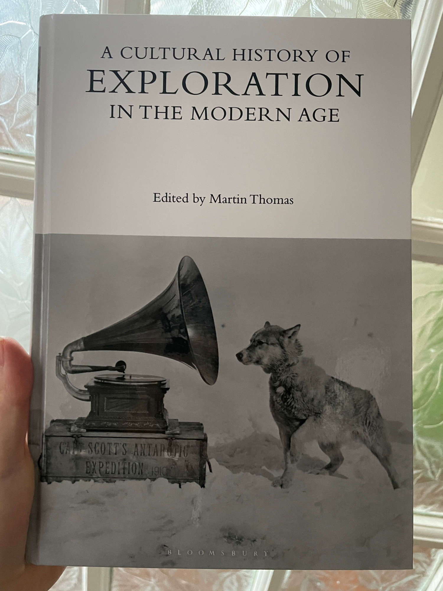 hand holdin a white book with a black and white photo of a husky dog in snow listening to a gramophone.  The title is "A cultural history of exploration in the modern age" edited by Martin Thomas