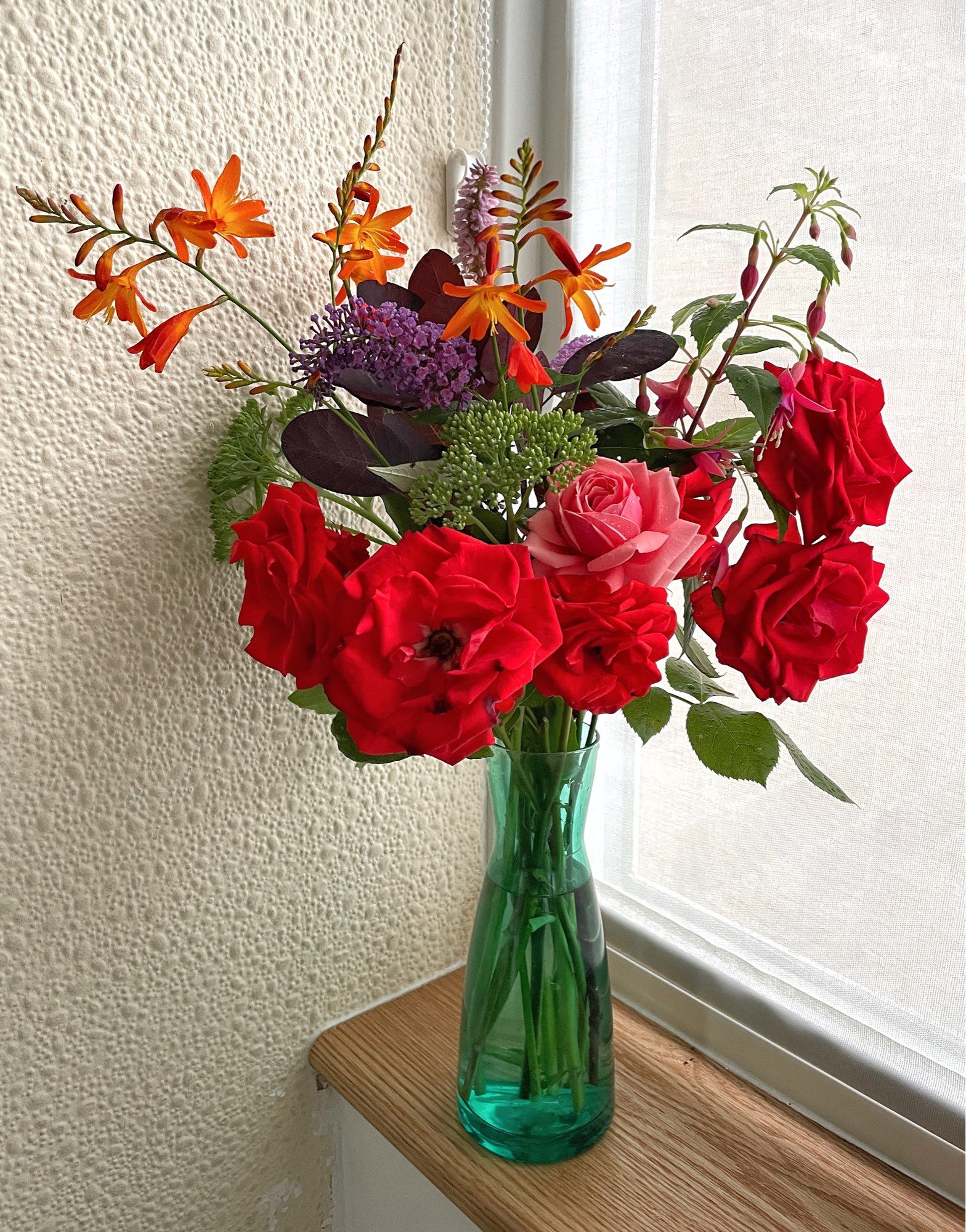 A large flower arrangement in a green glass vase. It stands in a wooden windowsill against a shaded window and pale wall. Red and pink roses, purple spikes, orange crocosmia, pink fuschia and foliage.