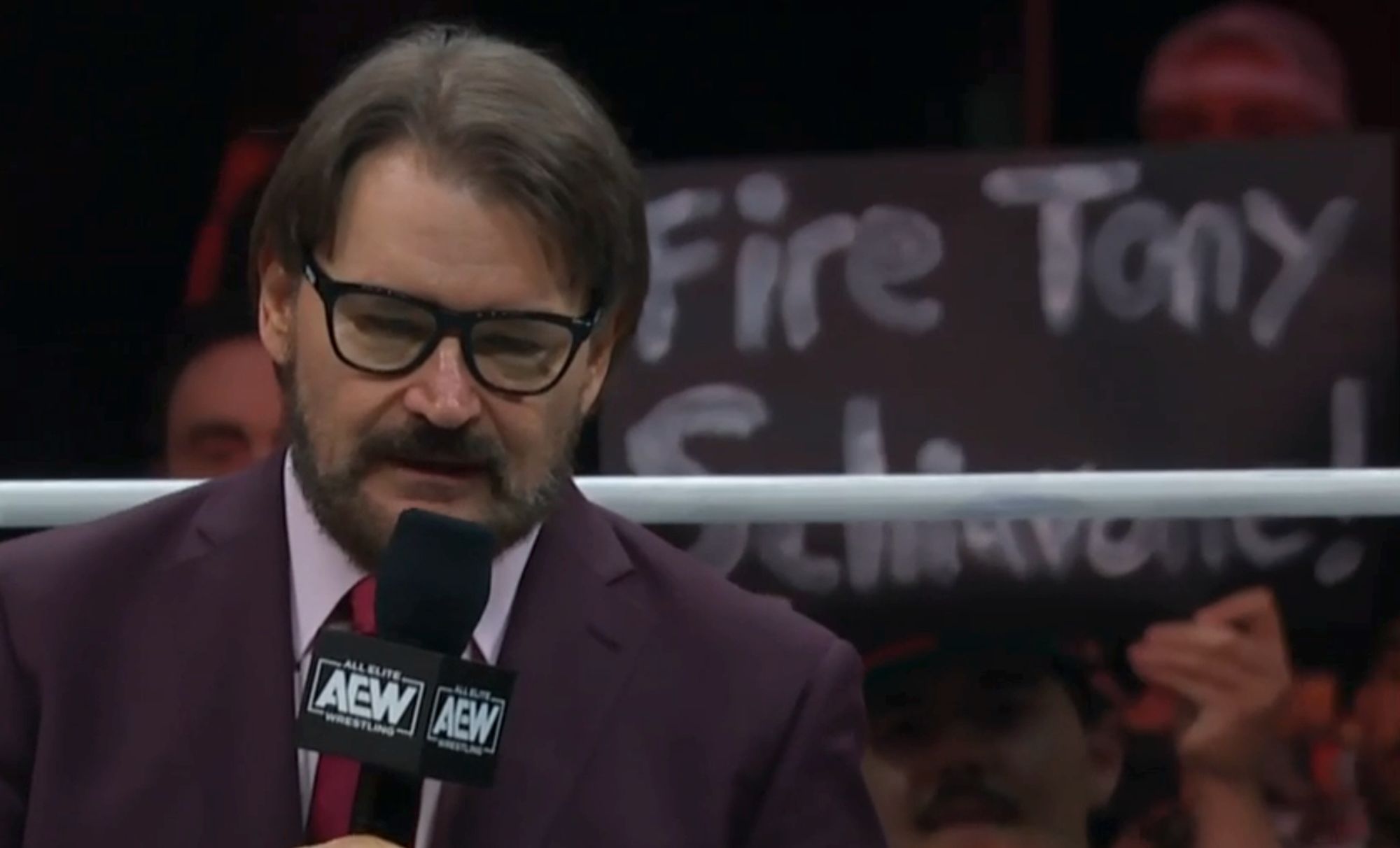 Tony Schiavone in the ring with a mic and a sign right behind him says "Fire Tony Schiavone!"