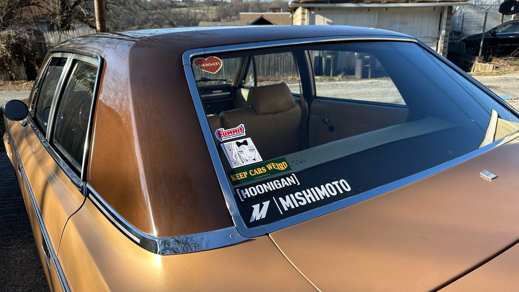 Photo of the rear glass of my 1969 AMC Rebel showing the sticker collection. Top is an OHOAT! Glitter Heart sticker, further down is Summit Racing, a manga panel of Chika Fujiwara from Kaguya Sama: Love is War making a smug face, an OHOAT “Keep Cars Weird” slap, an OG Hoonigan censor bar, and a Mishimoto sticker.