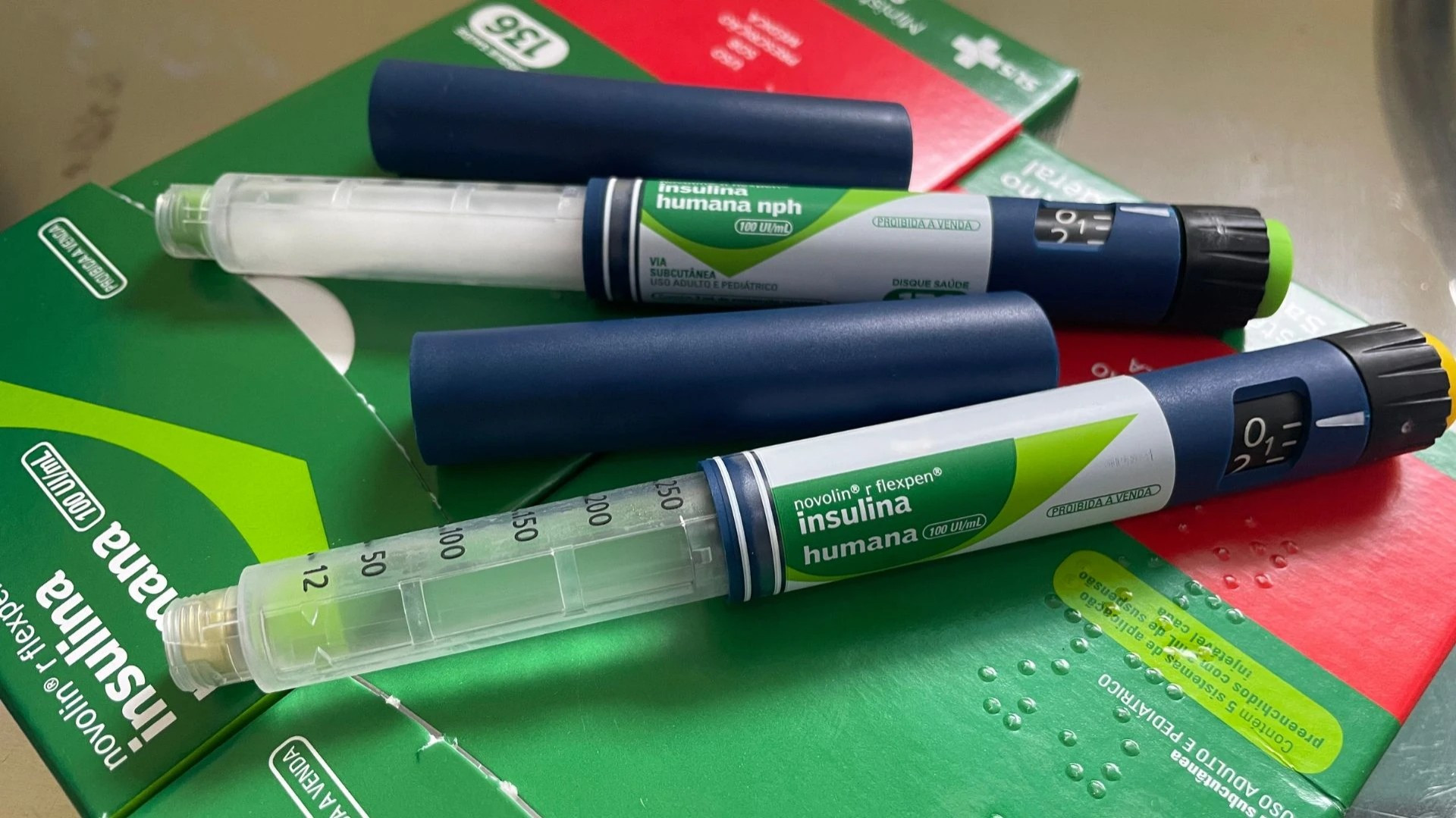 A imagem mostra um kit para o tratamento do diabetes. No centro, há uma caixa verde e branca com a palavra 'insulina' escrita em letras grandes e em negrito. Ao lado da caixa, estão três canetas injetoras de insulina. As canetas são cilíndricas, com corpo azul escuro e detalhes em verde. Uma das canetas está com a tampa removida, mostrando uma agulha fina e transparente na ponta. As canetas possuem um visor numérico que indica a dosagem de insulina a ser aplicada. O fundo da imagem é um pouco escuro, com algumas sombras. A marca da insulina é 'novolin r flexpen'. Este tipo de caneta é utilizado por pessoas com diabetes para administrar a insulina necessária para controlar os níveis de açúcar no sangue.