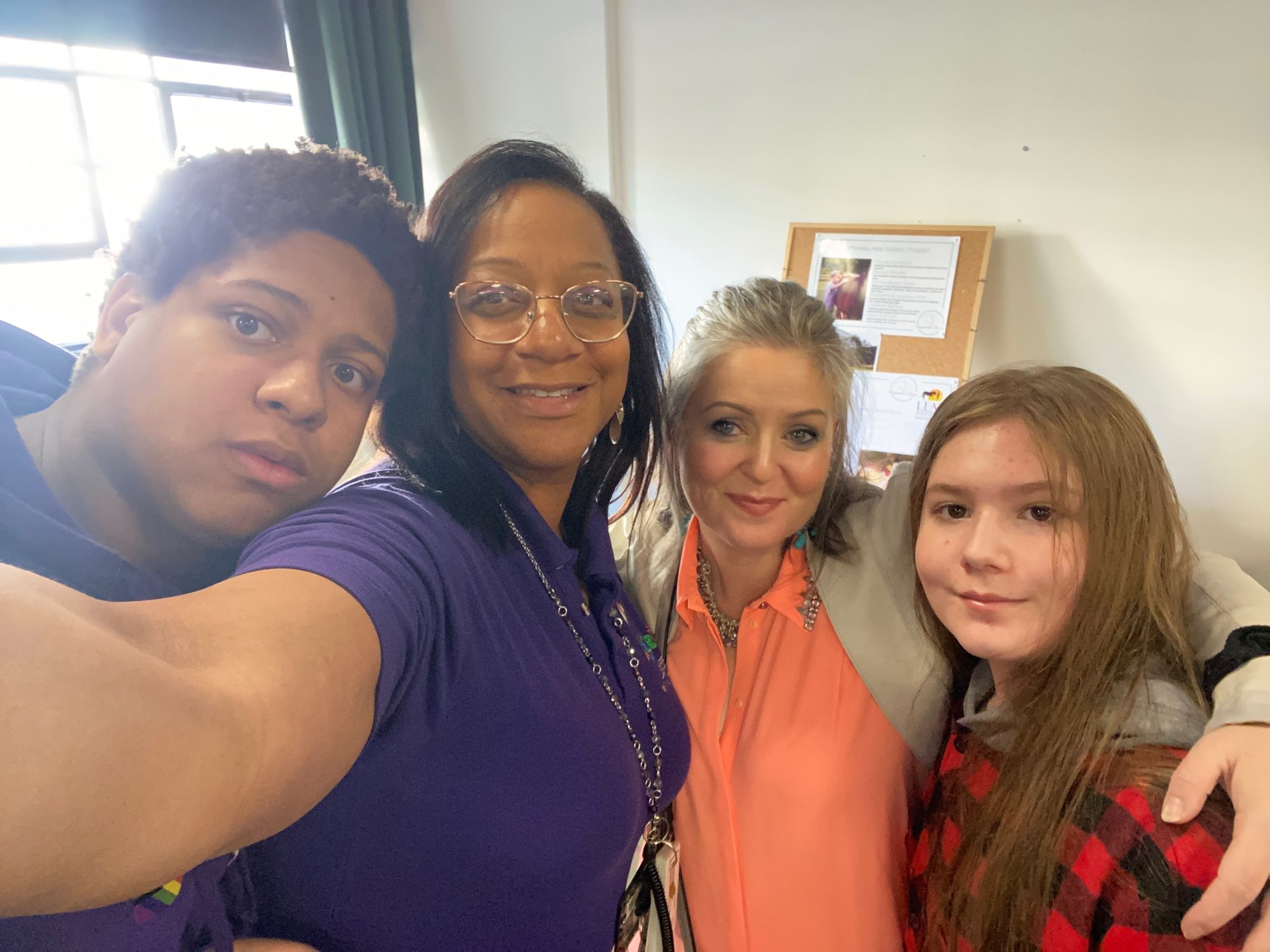 A group of four people from left to right a young black teenager, black adult woman who has black hair just on her shoulders a purple polo shirt and gold rimmed glasses. She is smiling and she is holding the camera then it is myself a short, middle-aged white woman with green mid-brown hair and blue eyes and wearing a coral pink blouse, finally, my son, a 12-year-old white boy with waist length light brown hair and brown eyes. We are smiling. He is wearing a red tartan shirt.