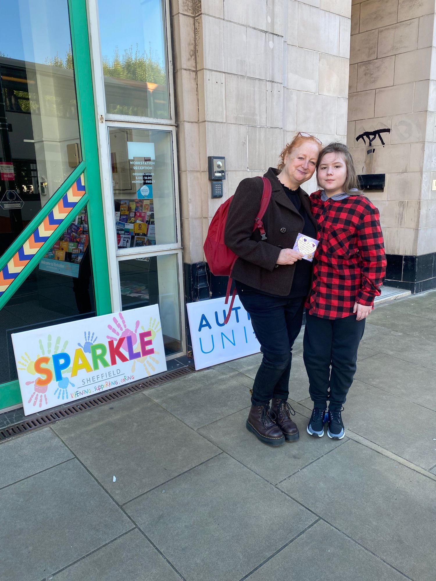 Outdoor photo of two people outside the workstation in Sheffield signs behind the two people read, sparkle Sheffield and autism union for two people in the photo from left to right of Chrissy , Meleady MBE, who is a middle-aged white woman with red natural curly hair that is tied back, she is dressed in black and white Doc Martens. She is holding a little heart and my son is the next to her. He is a 12-year-old white boy with very long light brown hair who has a plaid red and black shirt and black trousers and trainers.