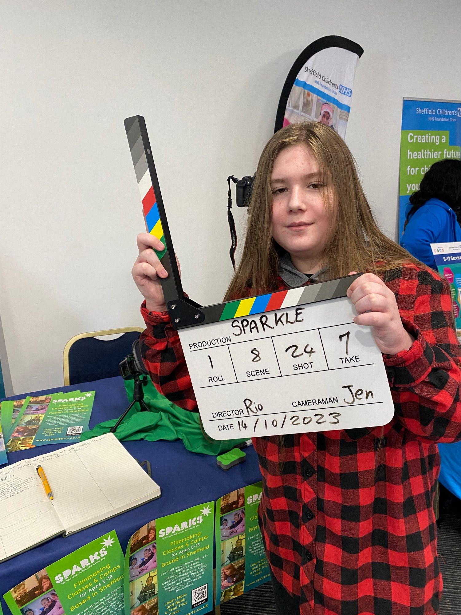 A white 12 year old boy with long light brown hair and 
with black and red plaid shirt holds a film clapper board , which says: production sparkle roll one scene eight shut 24 take seven director, Rio cameraman Jen, date, 14th of October 2023 is smiling