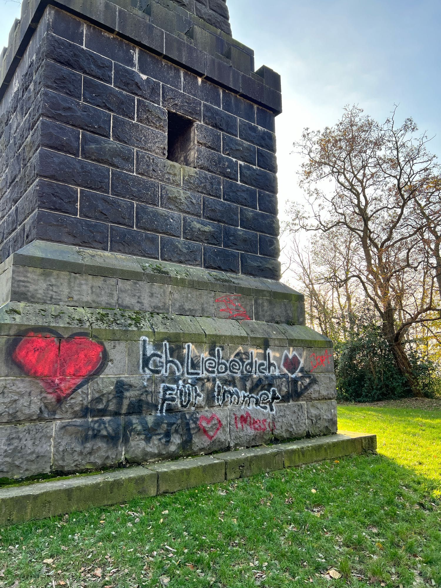 Das Bild zeigt den Sockel eines steinernen Denkmals oder Turms, der mit Graffiti versehen ist. Die Graffiti bestehen aus mehreren roten Herzen und einem deutschen Text. Die Hauptnachricht lautet “Ich liebe dich für immer”. Zusätzlich gibt es weitere rote Schriftzüge und Markierungen um den Haupttext herum, darunter ein kleineres rotes Herz mit den Worten “I miss u”.

Die Steinstruktur scheint Teil eines größeren Denkmals zu sein, das aus dunklen Ziegeln oder Blöcken besteht. Der Sockel ist quadratisch, und die Umgebung ist grasbewachsen. Im Hintergrund sind Bäume und ein klarer Himmel zu sehen, was darauf hindeutet, dass es sich um eine Außenaufnahme in einem Park oder einem historischen Gebiet handelt.