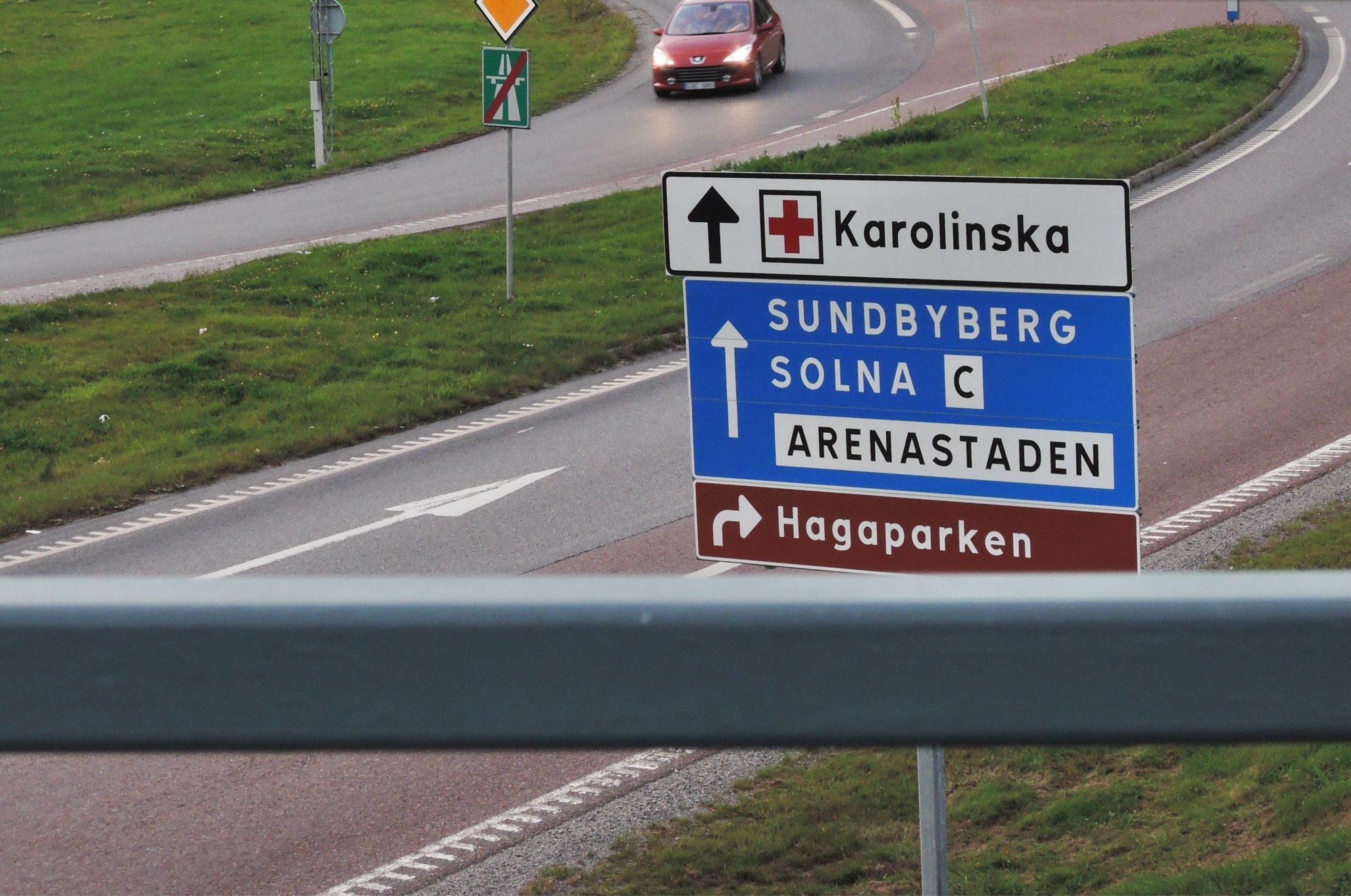 Verkehrssituation, von oben auf die Ausfahrt einer Schnellstraße mit grüner Böschung fotografiert. Im Vordergrund eine Leitplanke, darunter eine vierspurige Straße, in der Bildmitte ein Hinweisschild in Weiß, Blau und Rotbraun für die Richtungen. Darüber fährt ein roter PKW mit Scheinwerferlicht auf der Zubringerspur ins Bild.