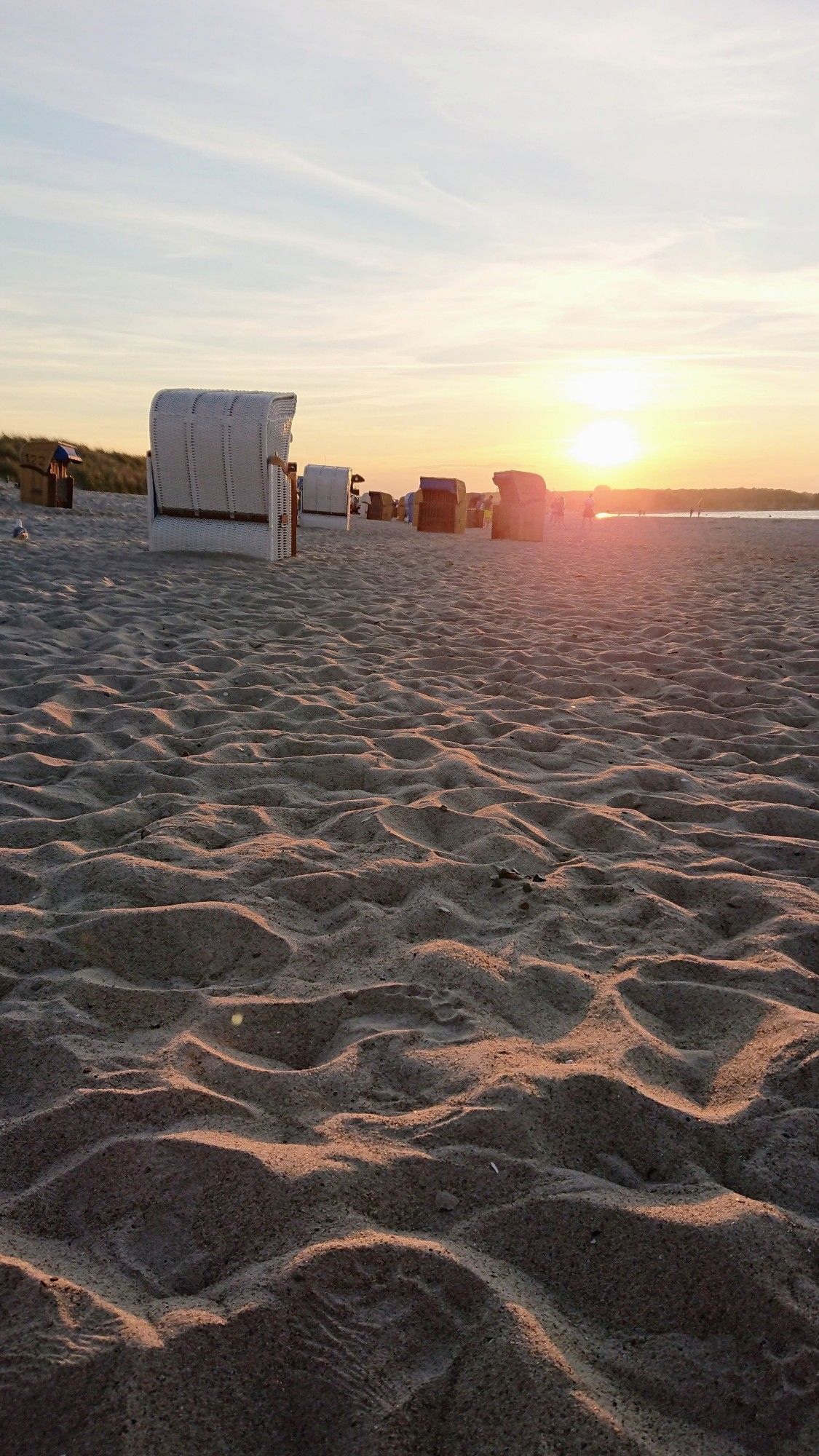 Ein Bild am Strand. Der Sand ist übersät mit Fußabdrücken. Strandkörbe stehen durcheinander. Die Sonne steht tief und färbt alles gelblich. In der Ferne sind Leute am Wasser