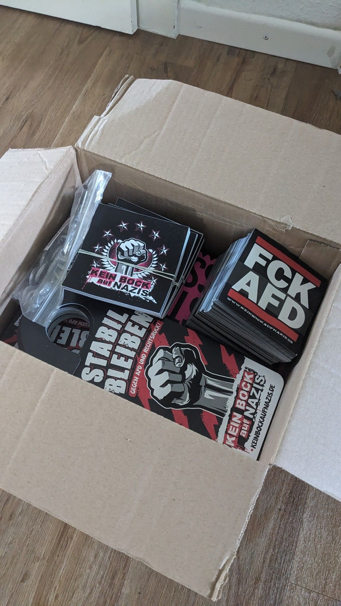 Foto eines geöffneten Lieferkartons.
Hierin sieht man verschiedene Antifa Artikel: FCK AFD Aufkleber, Kein Bock auf Nazis Aufkleber, Stabil bleiben - Kein Bock auf Nazis Türhänger, Kein Bock auf AFD Pins. Darunter (nicht/kaum sichtbar) befinden sich ca. 200 Flyer.