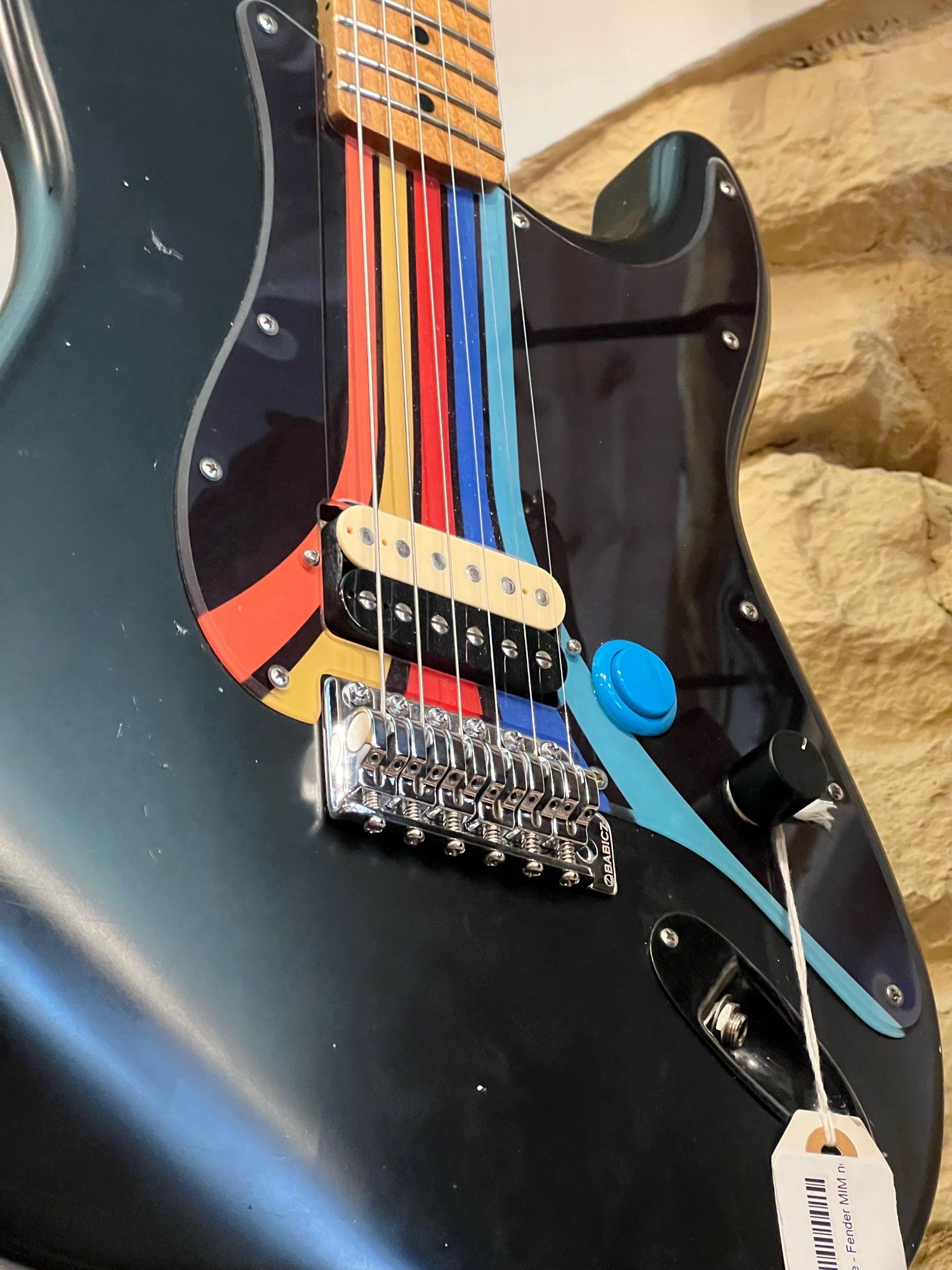 "Guitari," as Kelly put it. A MIM Fender Strat with a flat black finish, a single humbucker pickup, a single knob, and a blue arcade button of indeterminable function. The black pickguard has orange, yellow, red, blue, and light blue stripes the same width as the neck that splay out at the bottom and evoke the Atari logo