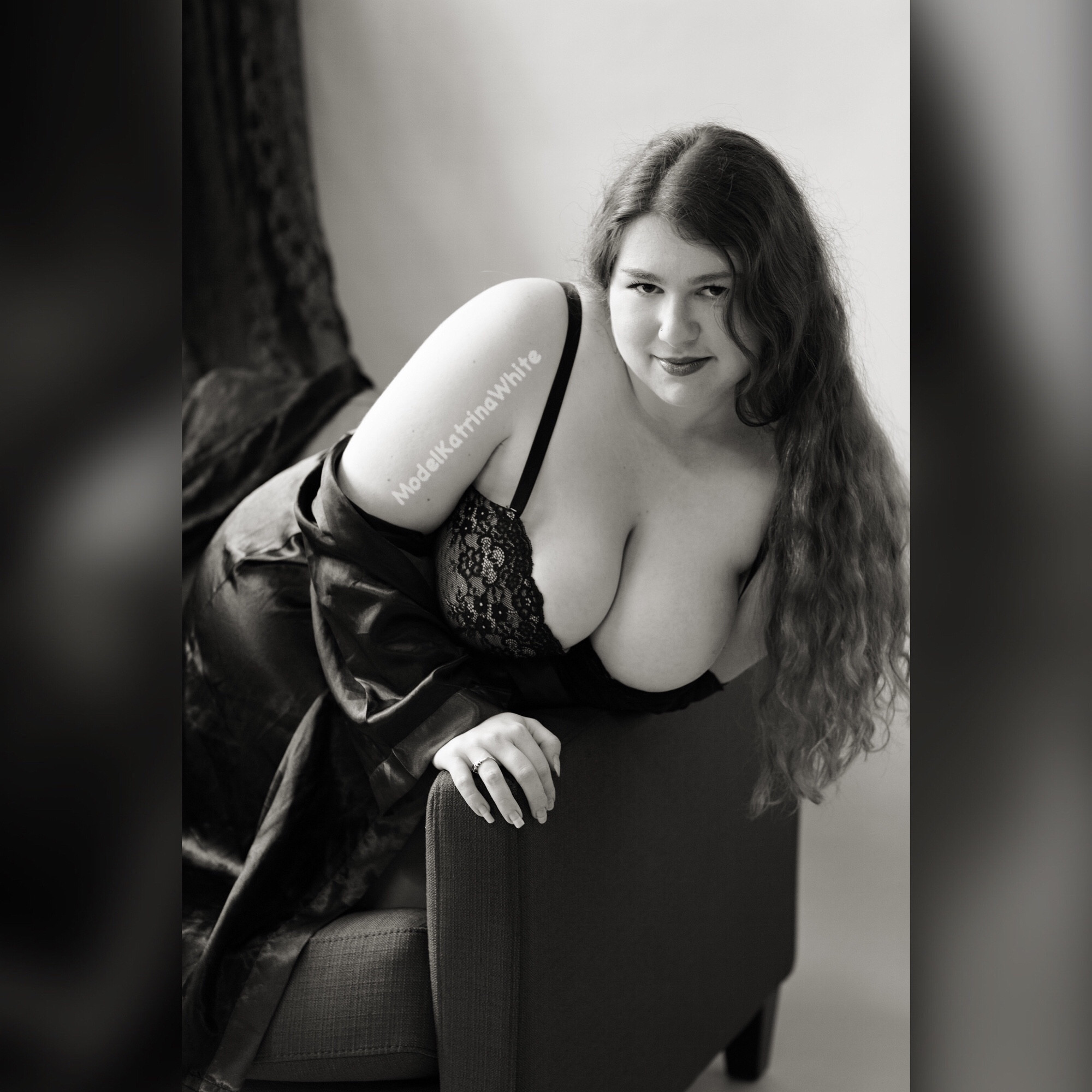 Black and white photo of Katrina white wearing lingerie. She leans over the side of a chair and her long wavy hair falls over her shoulder. Her shiny silk robe has slipped off her left shoulder and you can see ample cleavage spilling out of her lace bra. #nsfw 🧡🖤