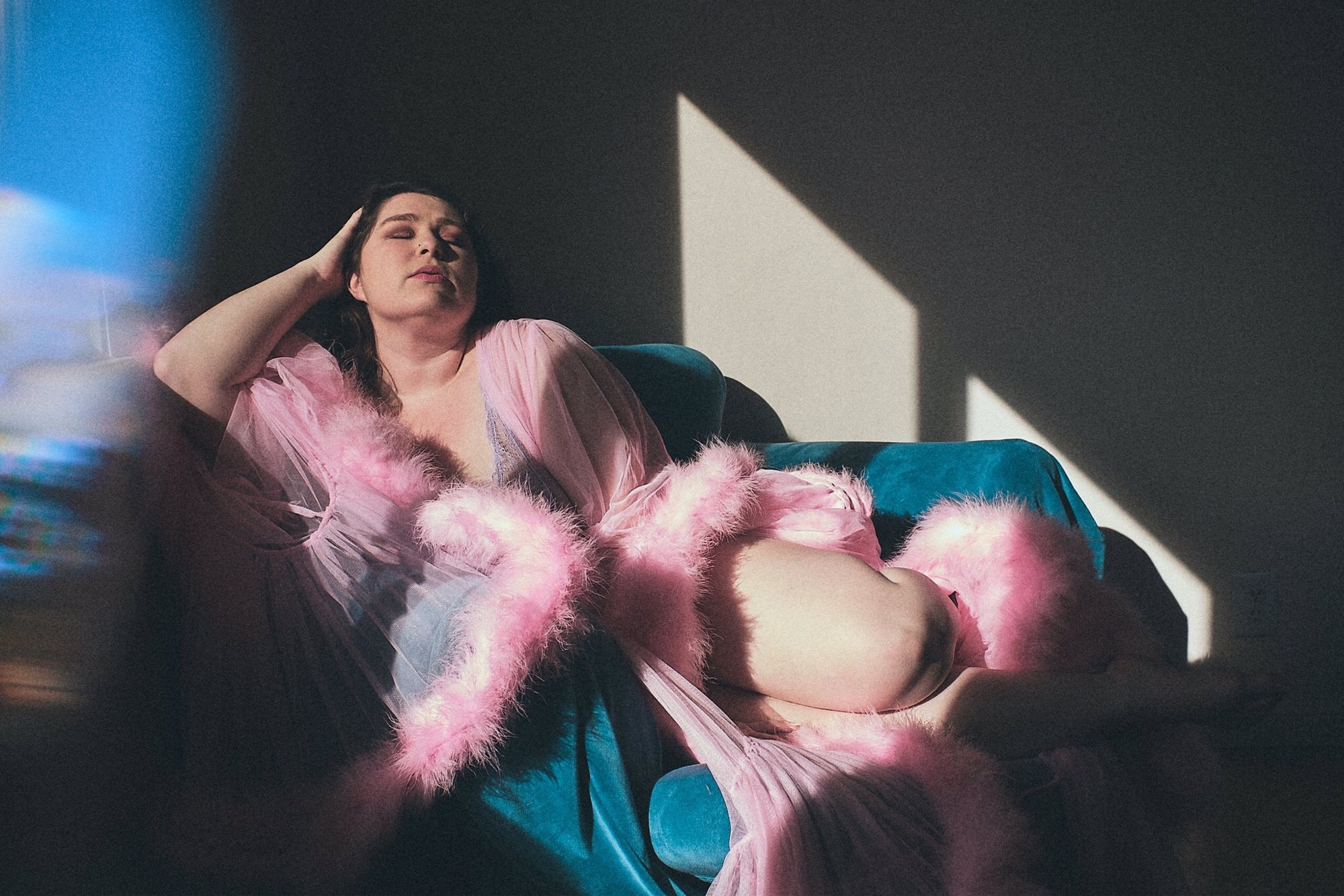 Katrina White lounges on a large blue velvet chair while wearing a sheer pink fluffy Catherine D’Lish robe. Her eyes are closed and the sun shining in the window creates shadows on the wall. #photography #art