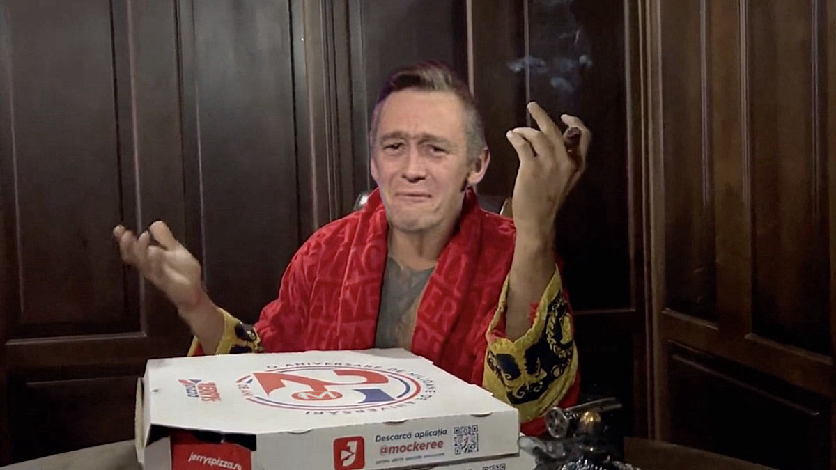 The Fast Show’s Ron Manager wearing a gaudy dressing gown and gesticulating theatrically with a cigar behind a telltale box of Romanian pizza