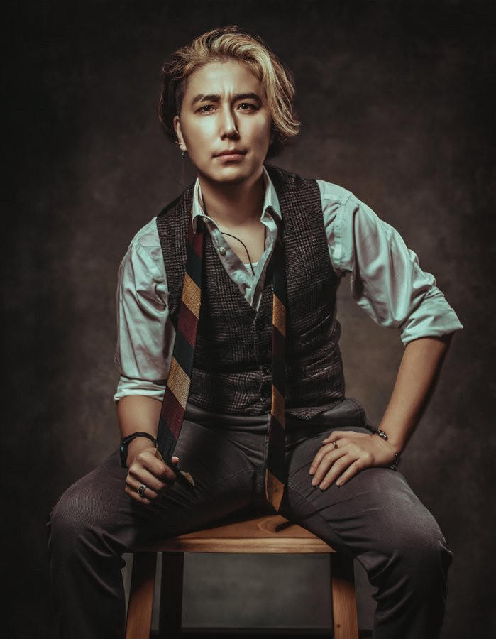 A handsome person sitting for a posed picture. It is Cai K an Asian person wearing a masculine suit. 