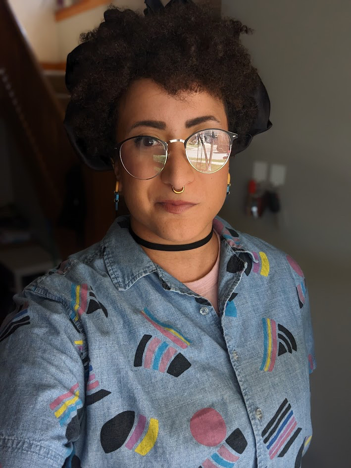 A picture of Bee Zelda, that shows a BIPOC person wearing glasses and a denim jacket. 