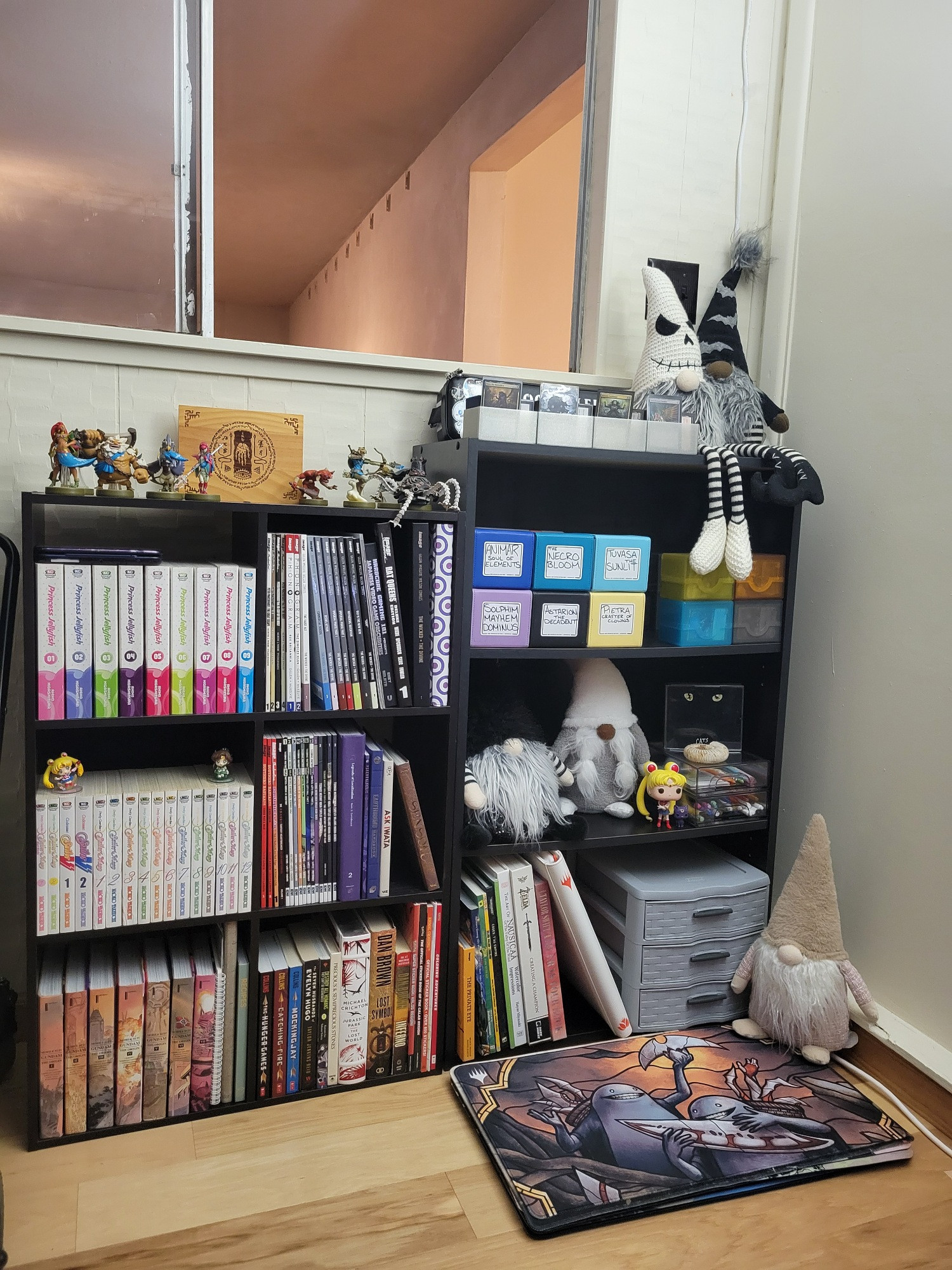 Two small, black wooden bookshelves sit cozily in the corner below an opened window. They're adorned with books, manga, deck boxes full of Magic the Gathering cards, figurines from Sailor Moon, and amiibo for Legend of Zelda Breath of the Wild, and gnomes. On the floor in front of the right bookshelf lays a stack of playmats.