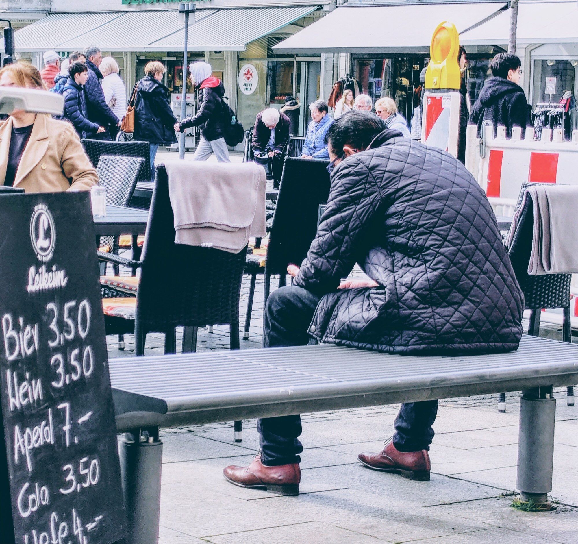 In der belebten Innenstadt, links vorne im Bild eine Preistafel eines Straßencafés, dahinter Stühle und Tische, teilweise mit Gästen. Im Hintergrund Geschäfte, Passanten. Eine Baustellenleuchte. Im Vordergrund steht eine Sitzbank aus Stahl, sie hat keine Lehne. Sie schließt sich a die Getränke-Tafel an, gehört aber nicht zum Café. Auf der rechten Seite der Bank, die rechte Bildhälfte fast ausfüllend, sitzt ein Mann mit dem Rücken zur Kamera. Er trägt Jeans, braune Schuhe, eine anthrazitgraue wattierte Jacke und sitzt vornüber gebeugt, die Arme auf den Oberschenkeln abgelegt. Möglicherweise hat er ein Smartphone in der Hand. Er sitzt alleine da, mitten im Trubel; die vielen Menschen, die den Hintergrund ausfüllen, sind weit entfernt, wesentlich kleiner und alle auf den Beinen.