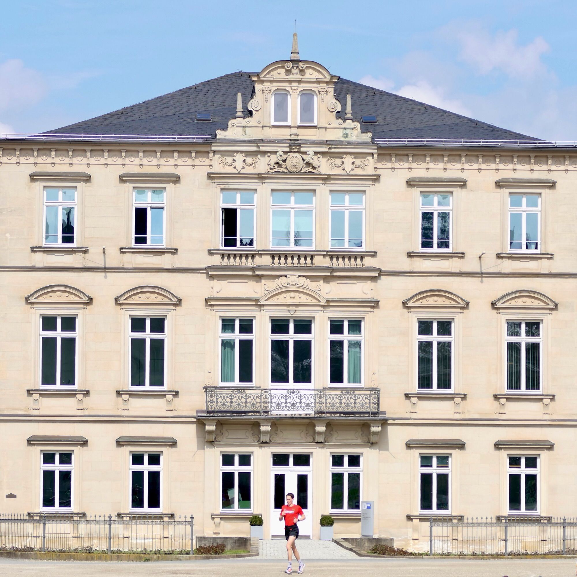 Ein repräsentatives historisches Gebäude im Stil der Neorenaissance, hell beige-gelb vor blassblauem leicht bewölktem Himmel; in der Mitte der symmetrischen Fassade, unter Ziergiebel und Balkon, befindet sich der Eingang mit weißer teilverglaster Tür. Genau vor der Mitte des Eingangs ist ein Jogger zu sehen. Er trägt ein rotes Hemd und schwarze Shorts, wirkt sehr dynamisch und entschlossen, wie er da so allein trainiert.