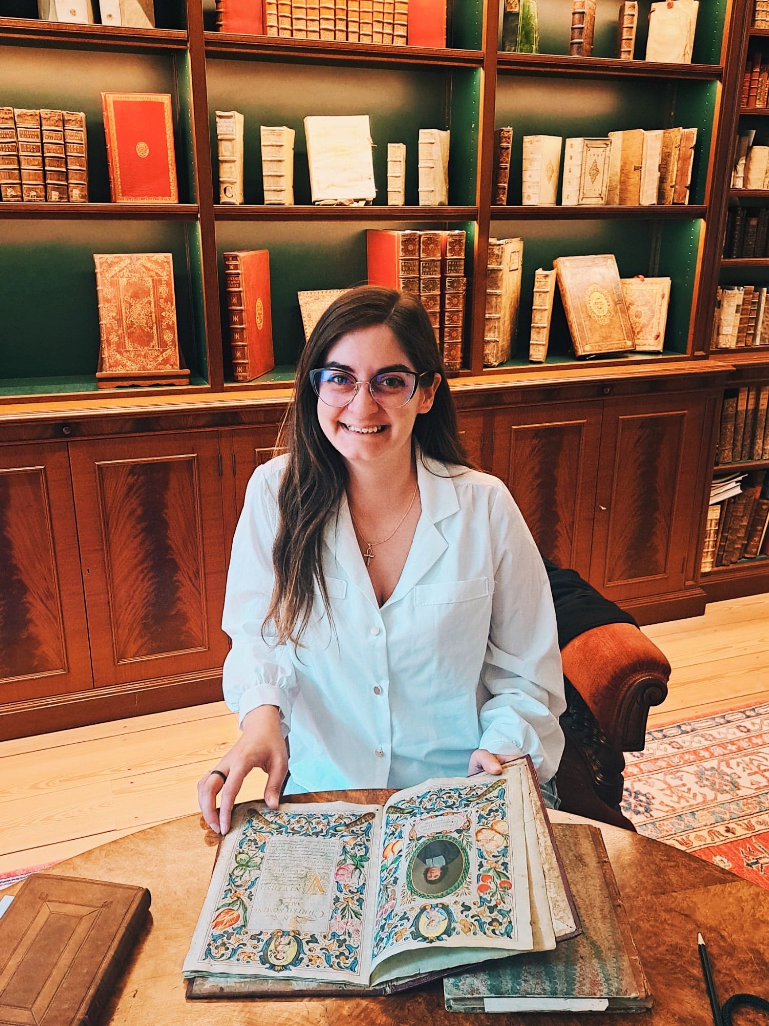 A woman sat at a wooden table with an antiquarian book in front of her, open to two colourfully illustrated pages