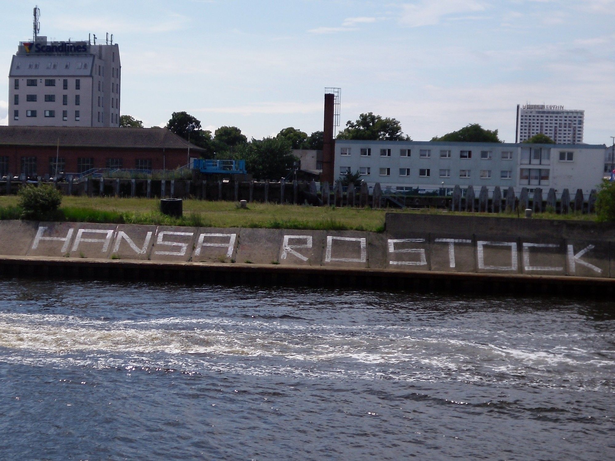 Der Schriftzug "HANSA ROSTOCK" auf einer Steinmauer. Das Foto wurde vom Wasser aufgenommen und am unteren Bildrand ist die Warnow zu sehen. Im Hintergrund befindet sich eine grüne Wiese mit Häusern dahinter.