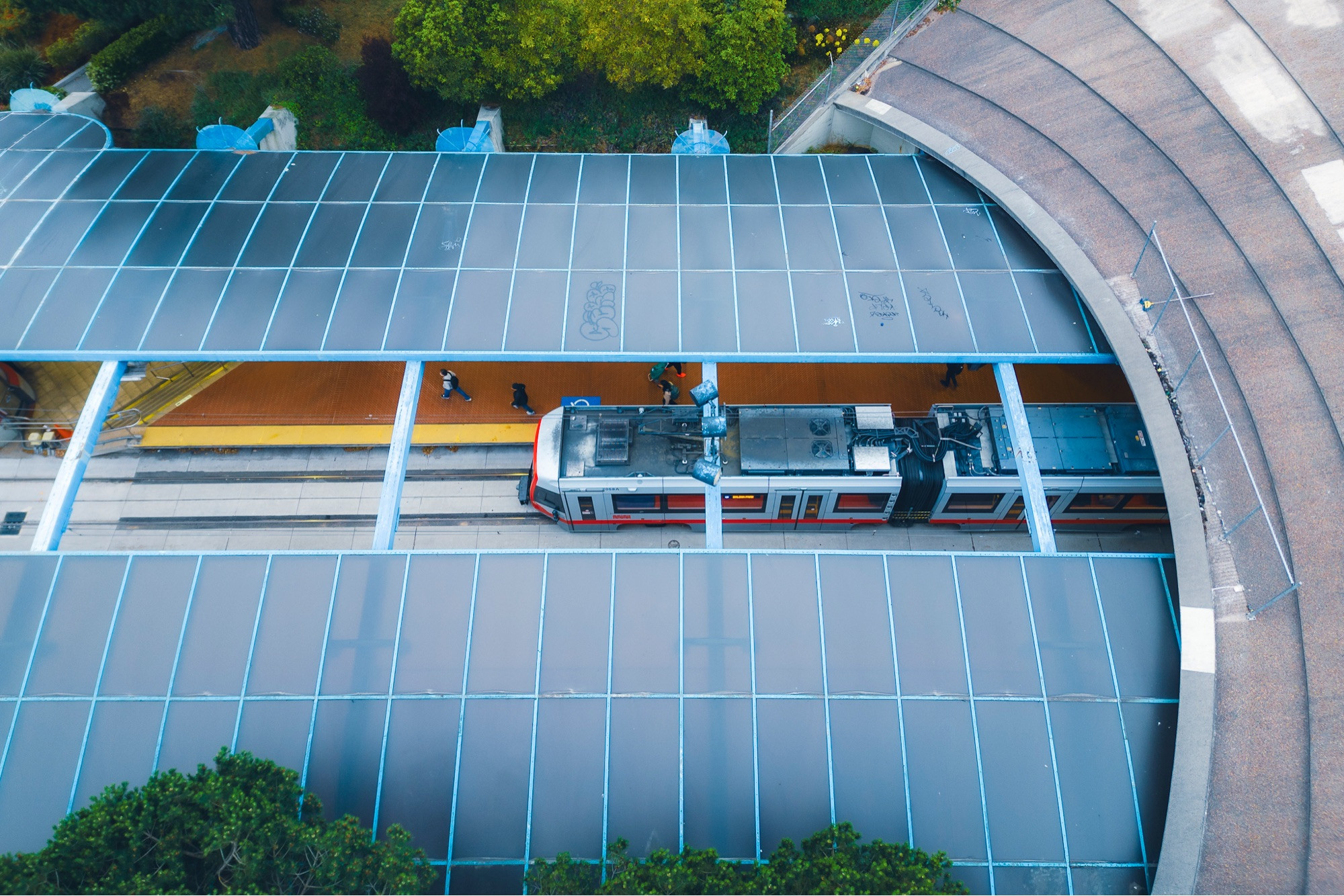 A train station seen from above, with a curved roof that has a gap in the center. The roof is made of dark black-blue reflective rectangular panels, with a light blue grid between them. Through the gap there is a train, and several passengers disembarking on the platform.