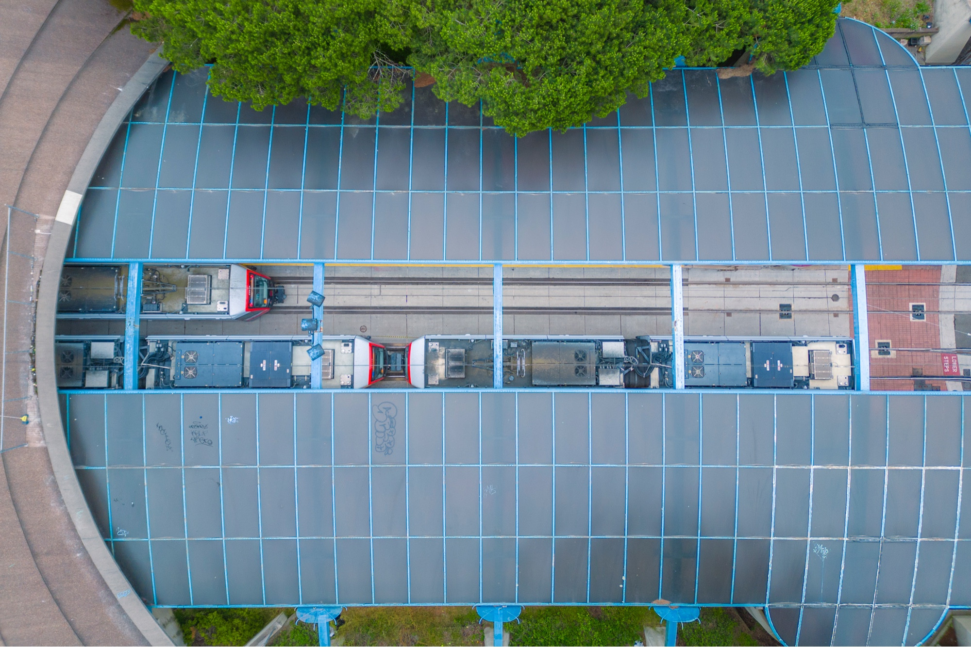 A direct overhead view of the station with two trains visible through the gap. There are trees and some grass visible beside the station.