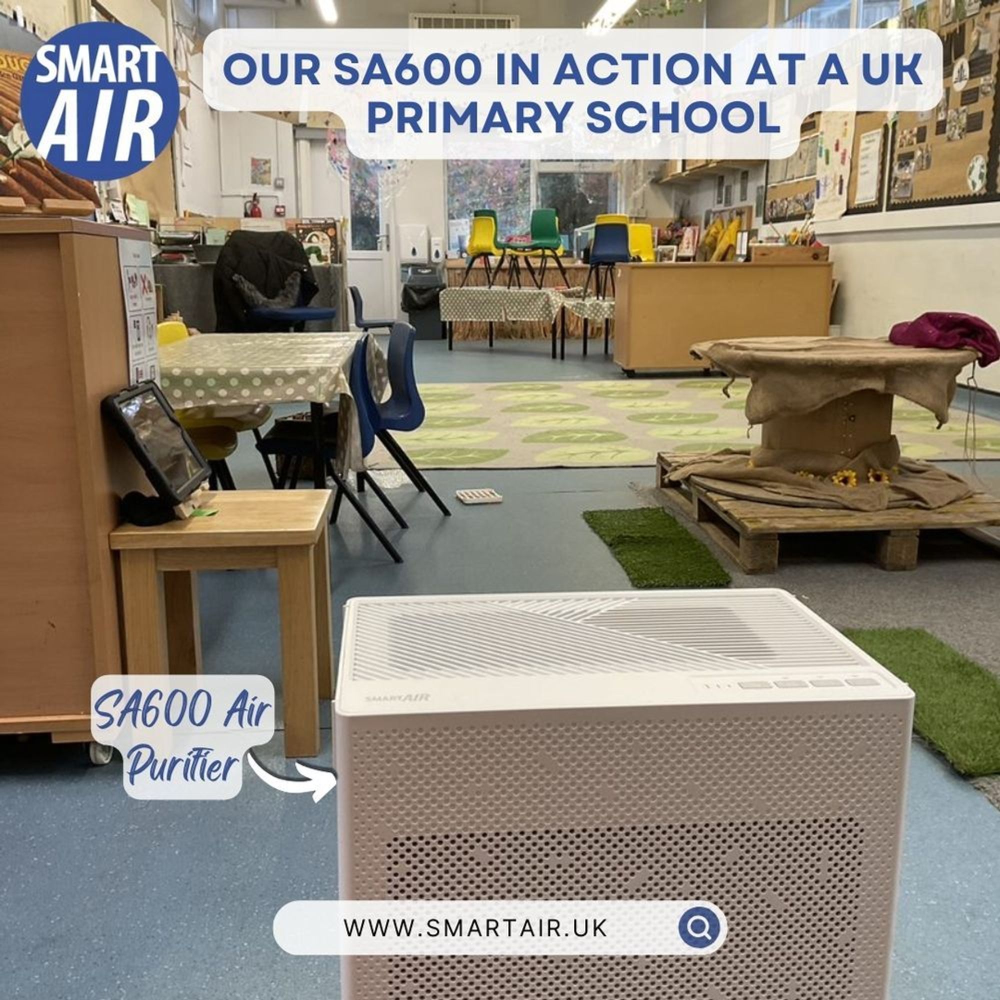 Smart Air SA600 air purifier placed in a UK primary school classroom. The image shows the air purifier on the floor in front of a child-friendly setup with colorful chairs, a round table, and educational displays on the wall. The room is bright and tidy, with the air purifier being the focal point.