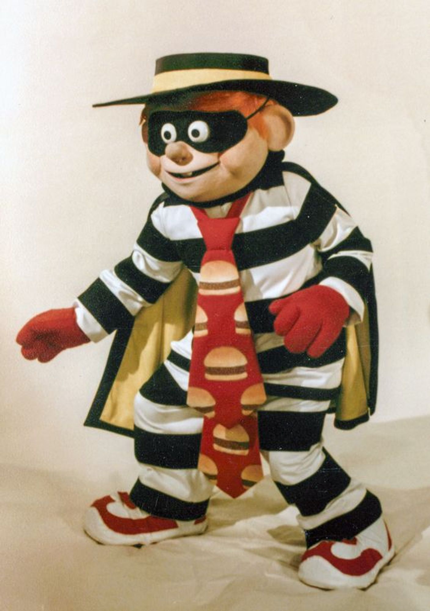 Promo photo of an actor in the costume for McDonald's McDonaldland "Hamburglar": a red-haired guy in a white-and-black striped jumpsuit (like a 1930s Hollywood stereotype of a prisoner), red-and-white sneakers, red gloves, a really big hat that's sort of a cross between a fedora, a porkpie, and a Cordoba, wearing a domino mask to hide his face and a very wide red tie with pictures of hamburgers on it. He's in a semicrouch like he's sneaking around.

Note: The character's signature phrase is "Robble, robble", used in varying intonations instead of regular speech.