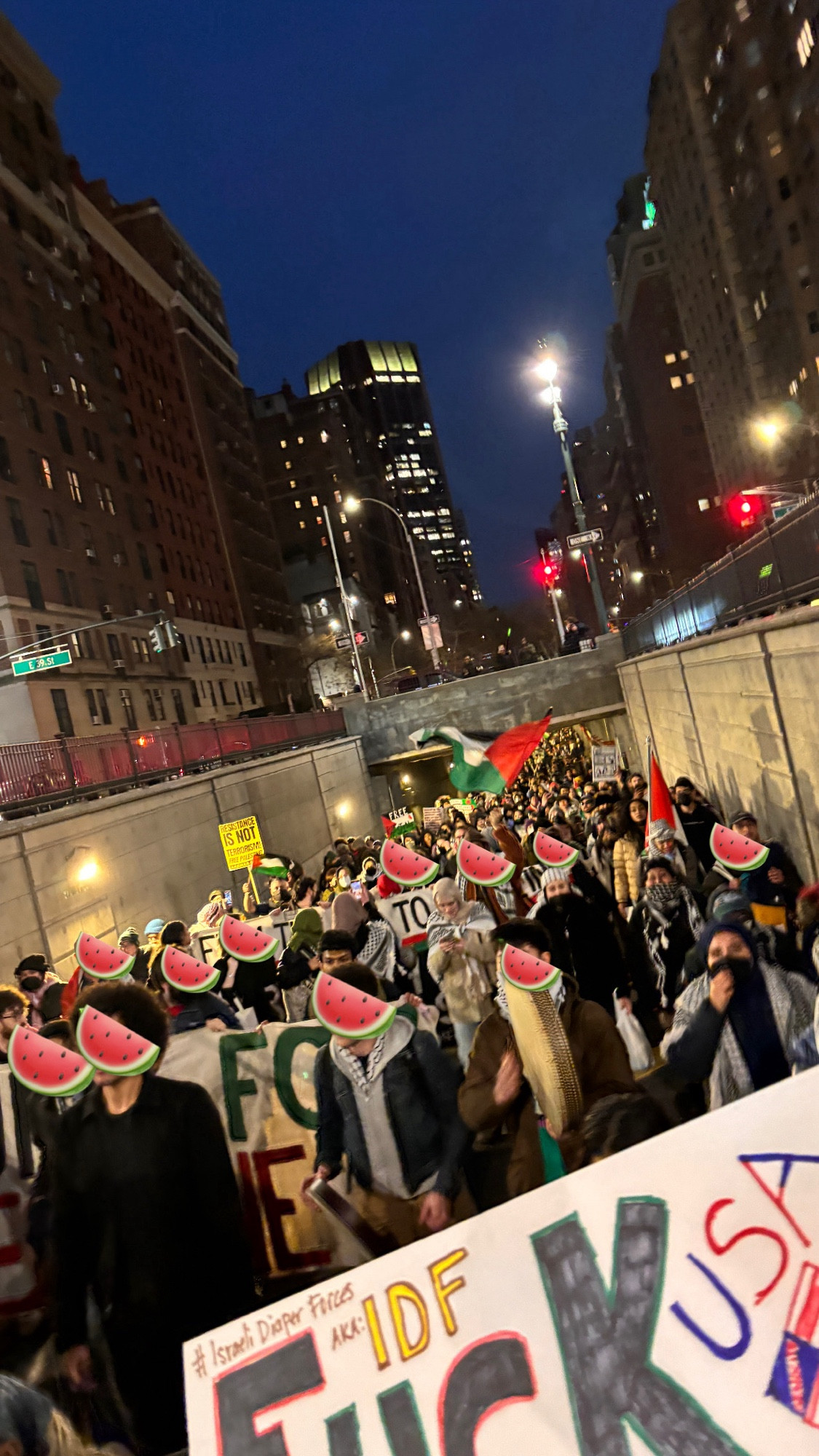 Thousands march for Palestine through the midtown tunnel with keffiyehs, Palestine flags, signs, and banners at night. Faces are obscured with watermelon emojis.